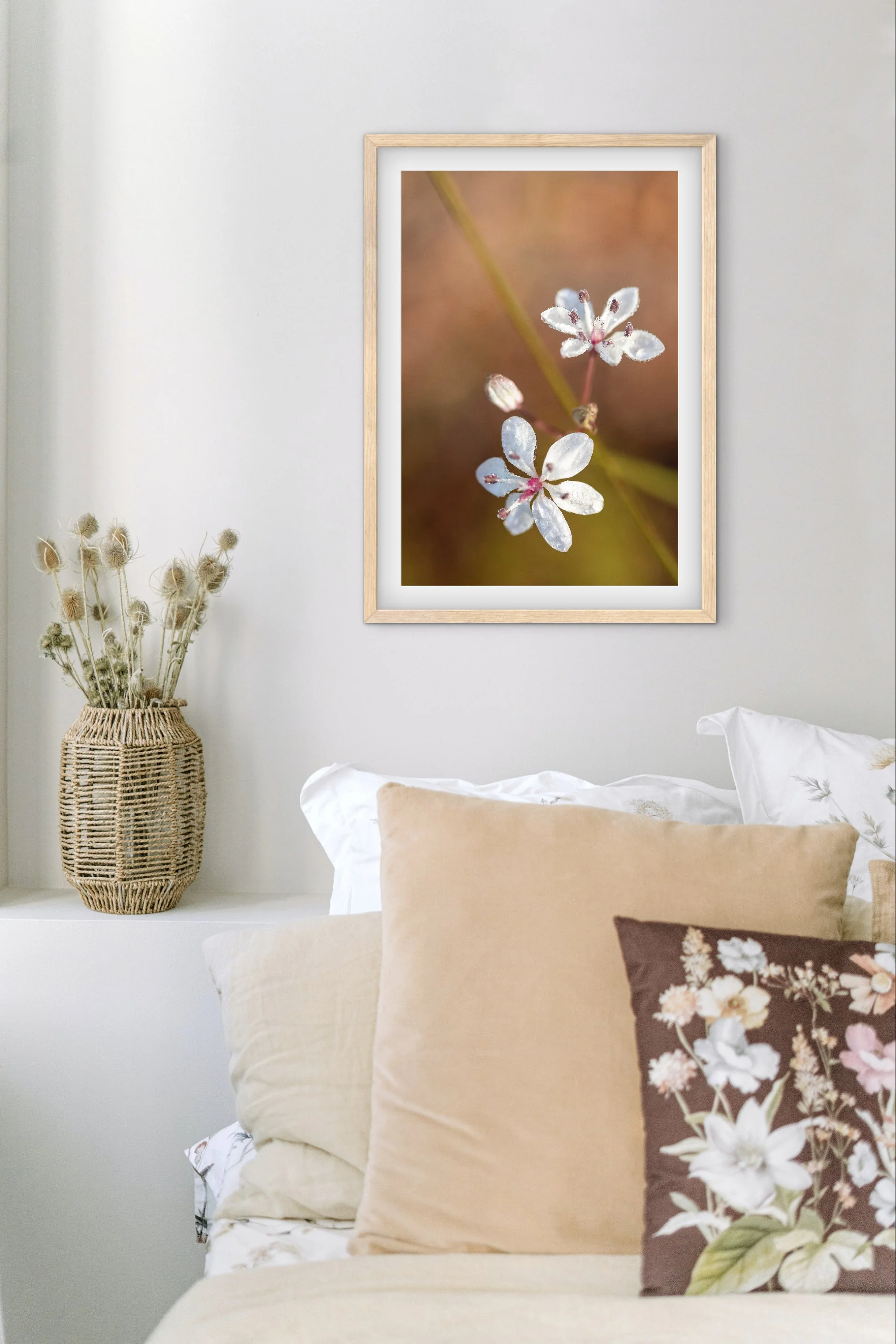 A bedroom corner featuring a framed macro photograph of white flowers with pink accents hanging on a white wall, next to a white shelf with a woven basket holding dried flowers, and a bed with beige, white, and floral pillows.