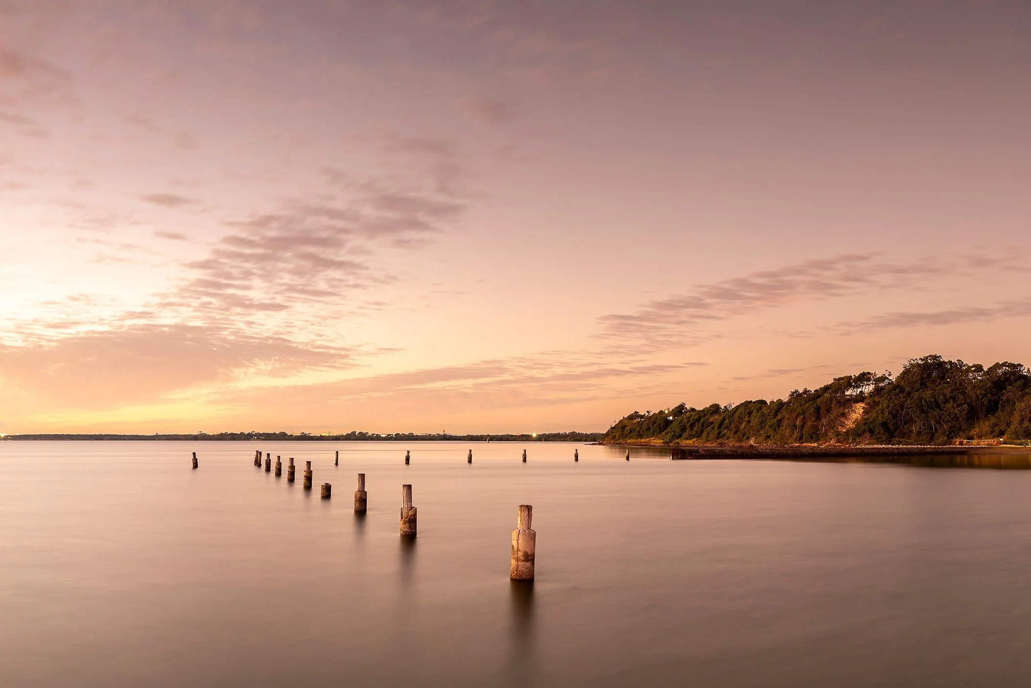 Old timber pylons extending into the calm waters of Bramble Bay at sunrise, with soft pastel skies and the Shorncliffe foreshore in Brisbane, Queensland.