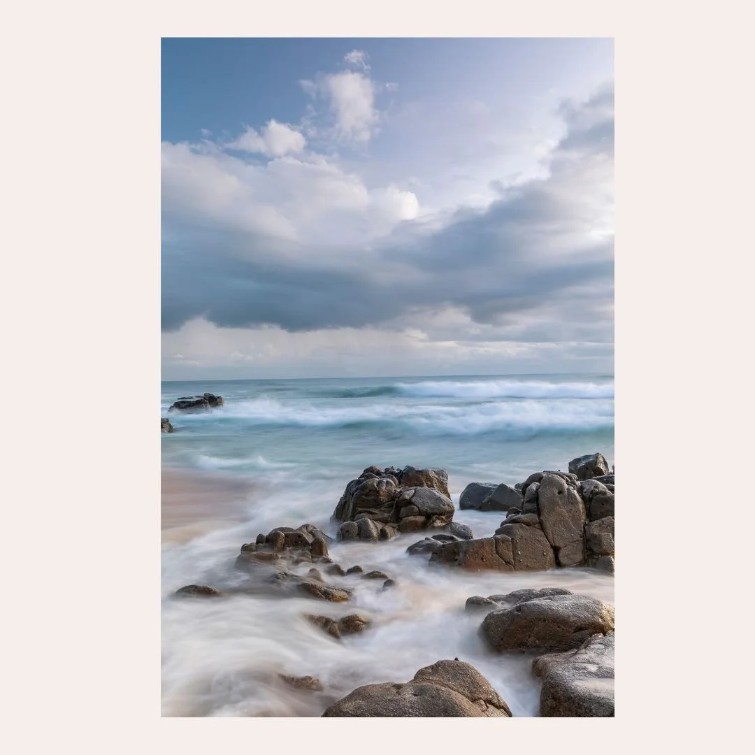 Long exposure photograph of waves flowing around coastal rocks at Coolum Beach on the Sunshine Coast, beneath dramatic clouds and patches of blue sky after sunrise.