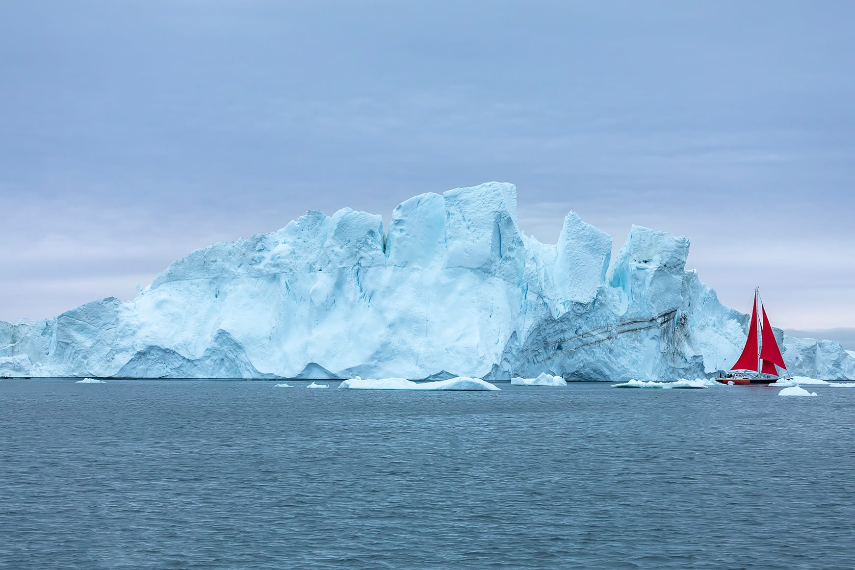 A massive iceberg in the waters near Ilulissat, Greenland, with a red-sailed yacht sailing alongside to show the scale of the ice formation.