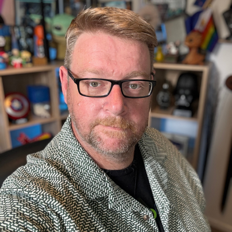 A smiling man with light brown hair and a beard, wearing a light purple button-up shirt, sitting outdoors against a stone wall.