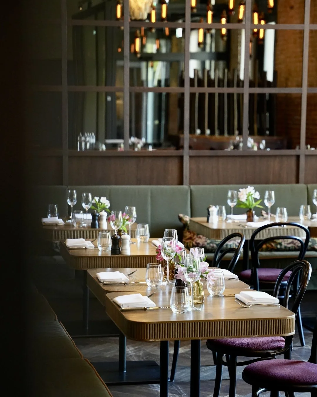 Ein elegant gedeckter Restauranttisch mit Blumen in Vasen, Gläsern und Besteck, im Hintergrund weitere Tische und eine Wand mit Holzlamellen.