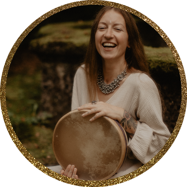 Eine lachende Frau mit dunkelblonden Haaren, die eine Ocean Drum hält und in der Natur vor einem Moos-bewachsenem Brunnen sitzt.