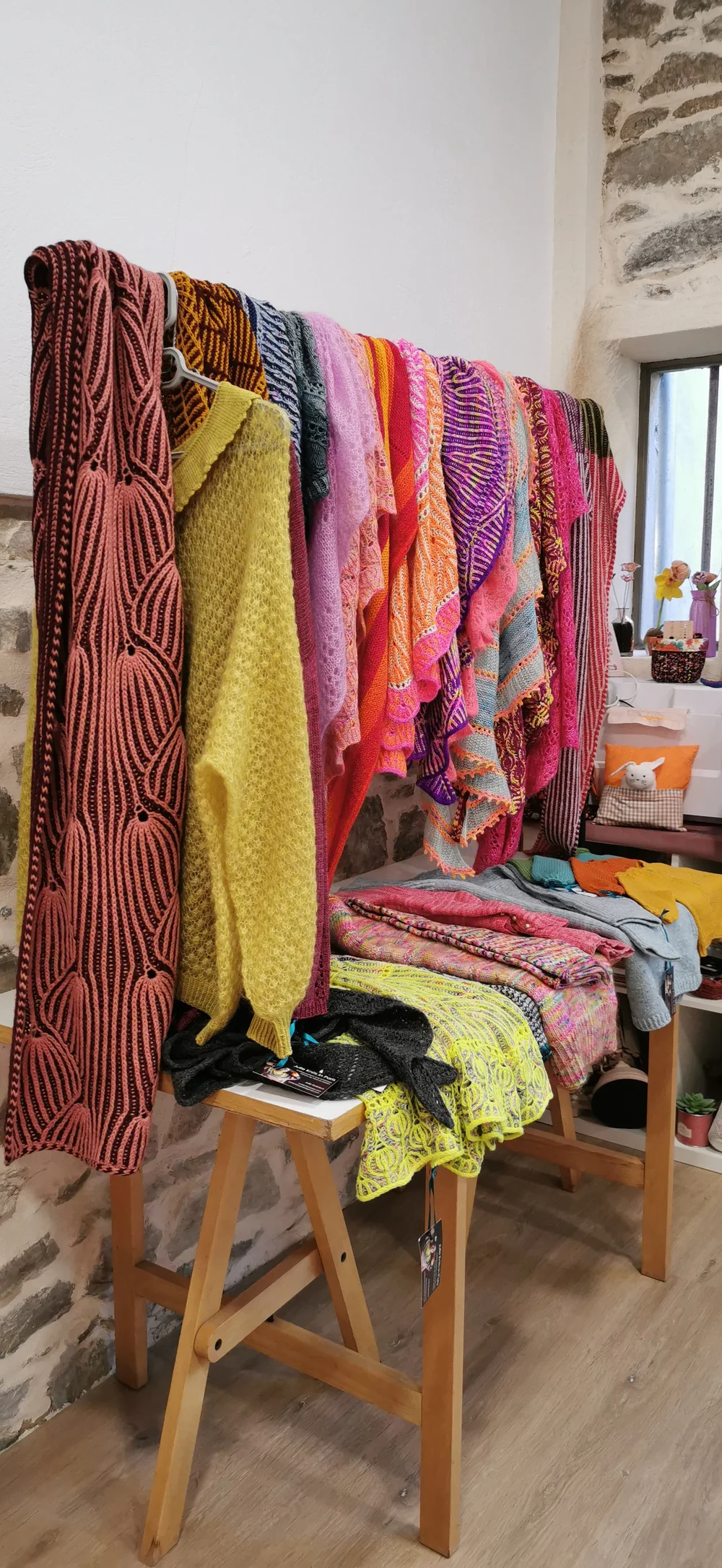 Brioche and colourwork shawls and knitwear hanging on a rack in a stone-walled room