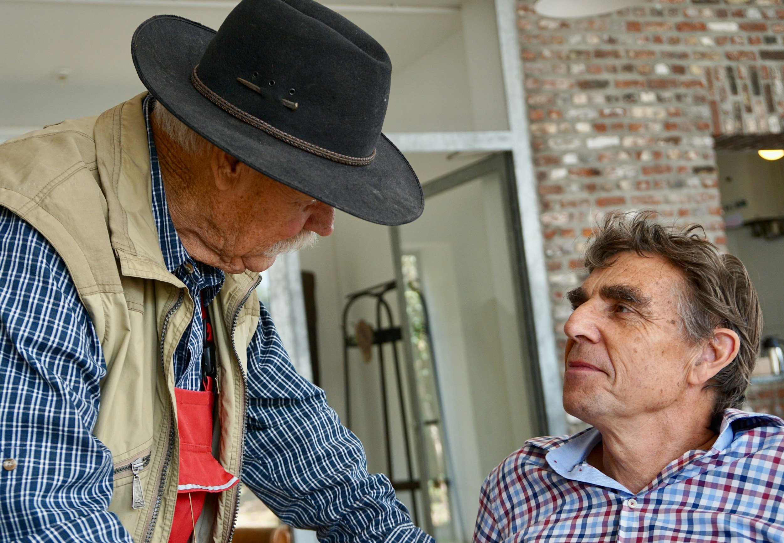 An elderly man with gray hair and a plaid shirt looking serious at a man wearing a wide-brimmed hat and a vest, inside a room with brick wall and glass door.