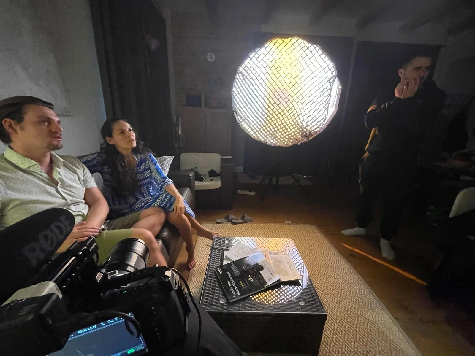 A film or photoshoot set with four people, two seated on a black couch and two standing. They are in a dimly lit room with a large circular light and photography equipment visible. One person is using a camera, and the others are observing or waiting