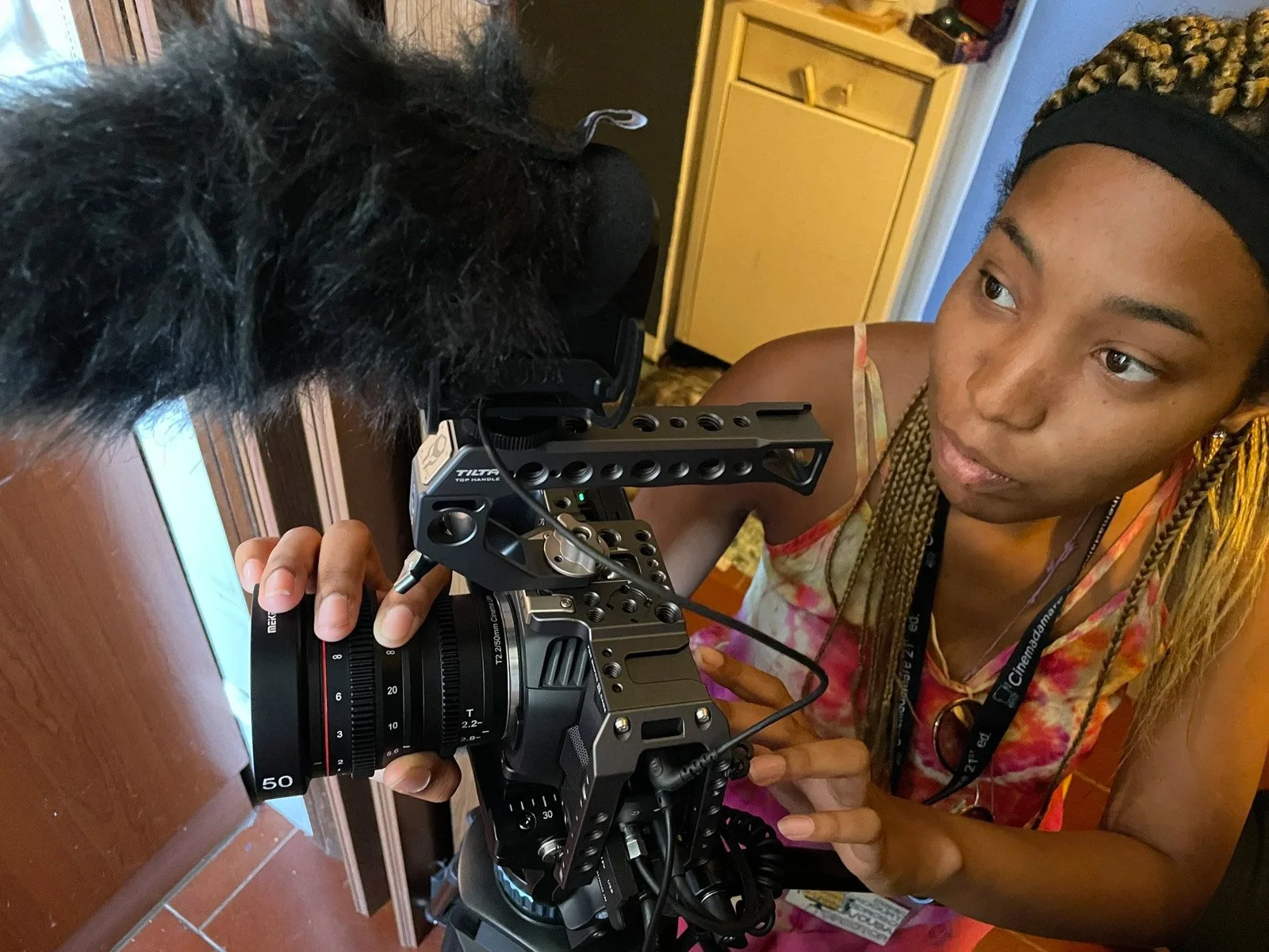 A woman with braids and a black headband is operating a professional video camera, filming a person with curly black hair near a wooden surface indoors.