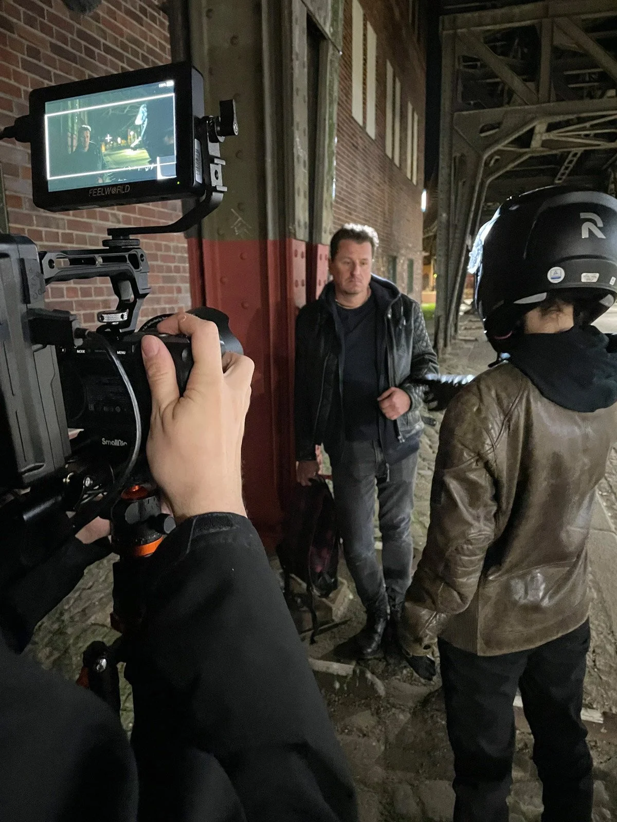 A man in a black leather jacket is being filmed by a camera operator. Another person wearing a motorcycle helmet and leather jacket is in the foreground. The scene appears to be shot outside under a structure with brick and metal elements.