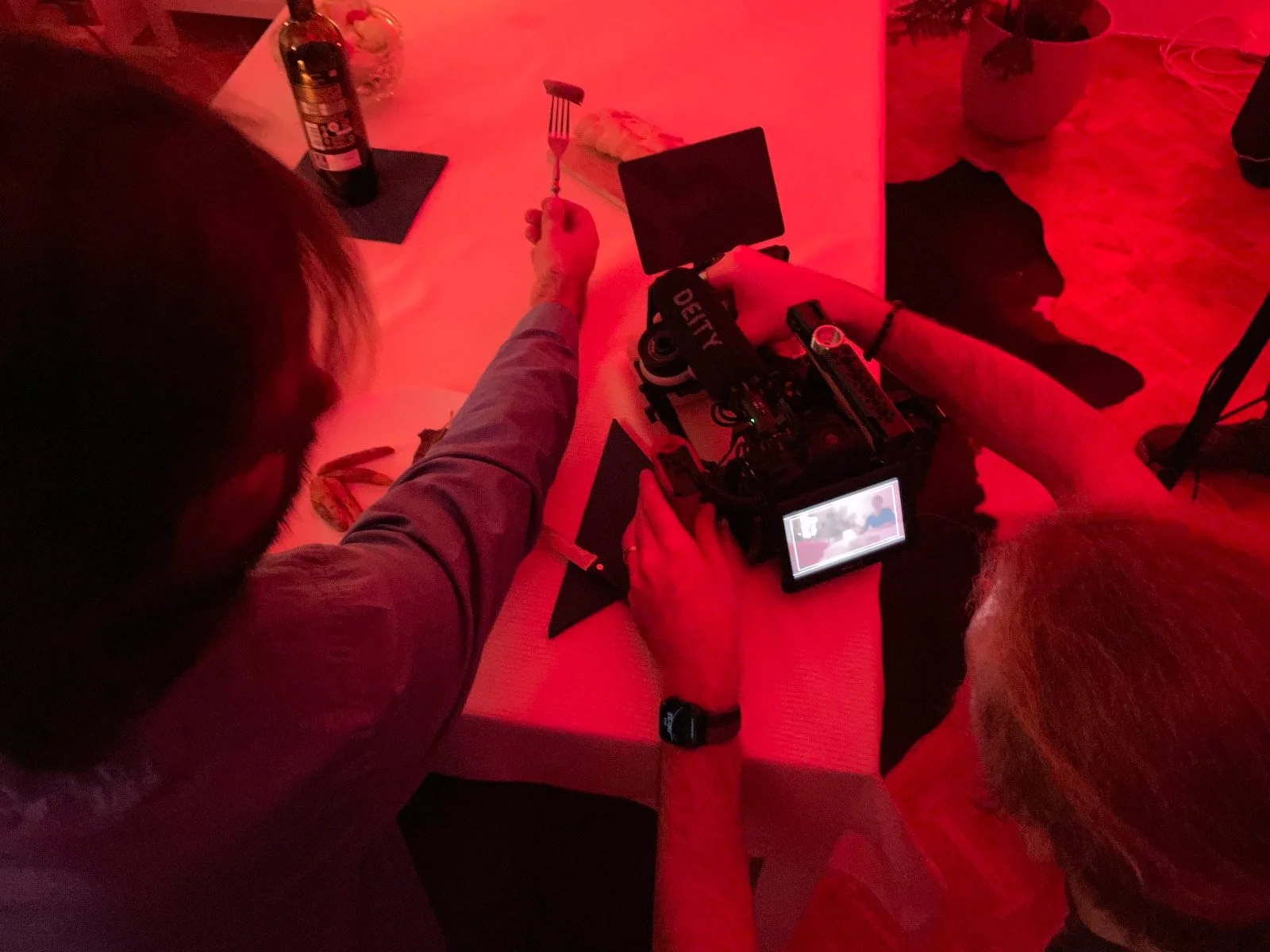 A person is sitting at a table with a camera and a red lighting environment. The person has a watch on their left wrist and is holding a camera with a screen showing a live view of the scene. There is another person beside them with gray hair, and so