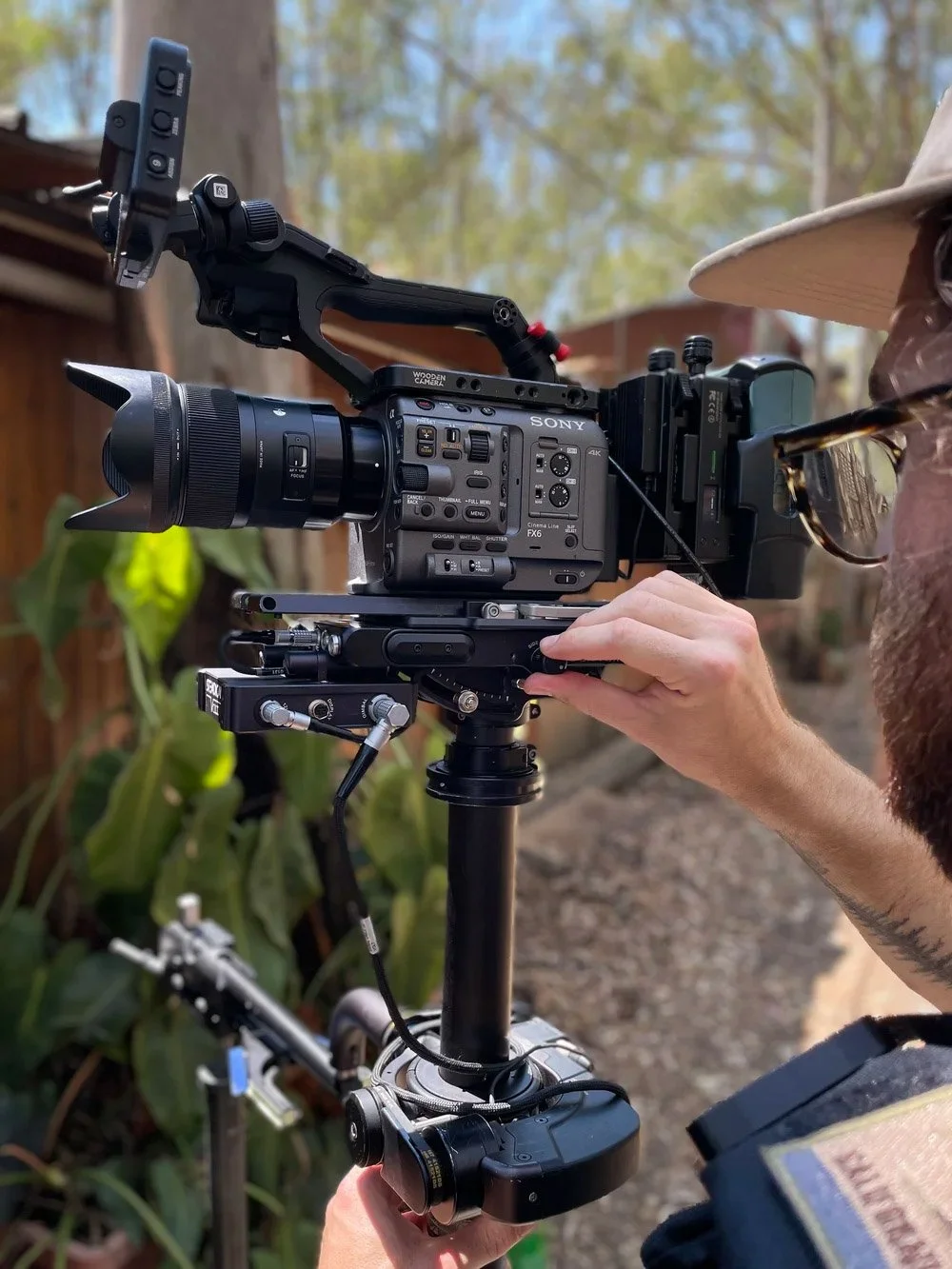 A person operating a professional Sony video camera mounted on a stabilizer in an outdoor setting with trees and a wooden fence in the background.