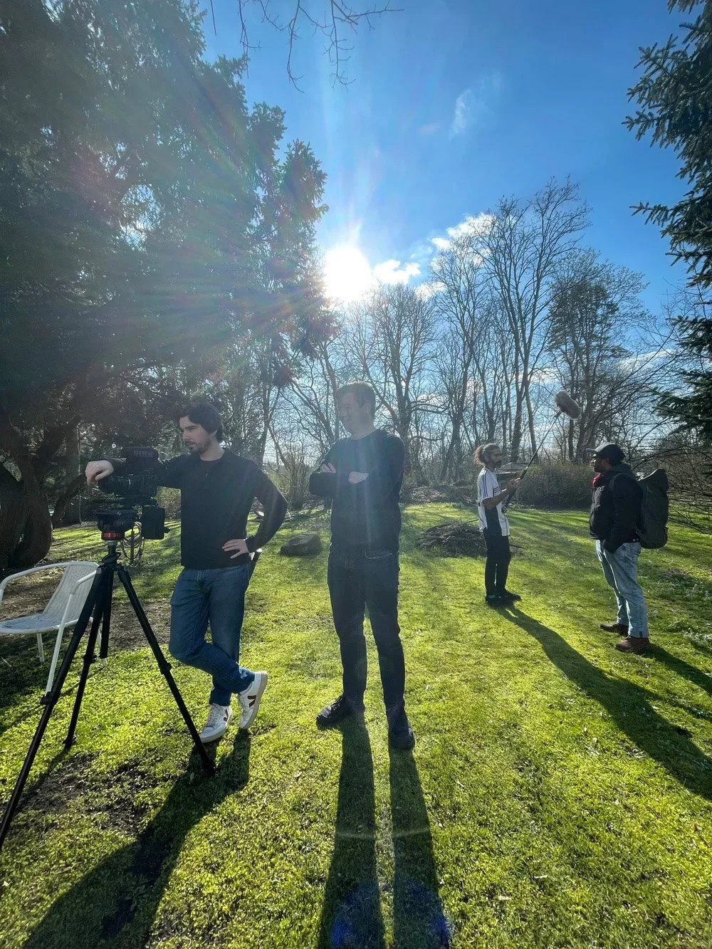 Group of people outdoors on a sunny day, one person operating a camera on a tripod, others standing on the grass, some holding recording equipment, with trees and a blue sky in the background.