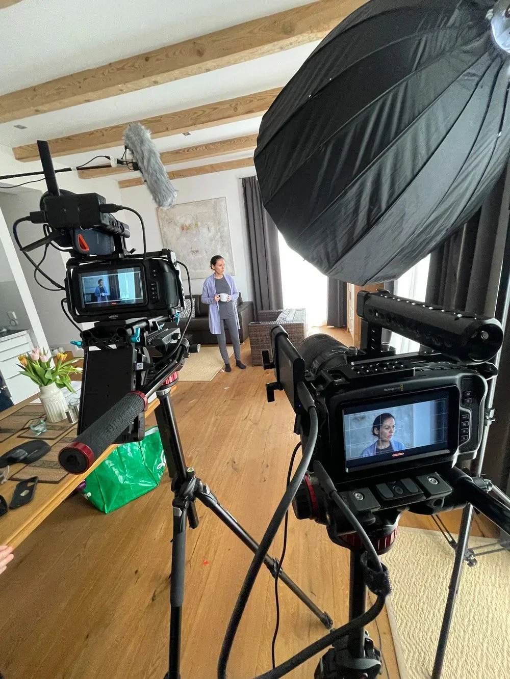 A woman standing in a living room, holding a mug, being filmed by two cameras with large softbox lights set up. The room has wooden floors, a bookshelf, and a window with curtains.