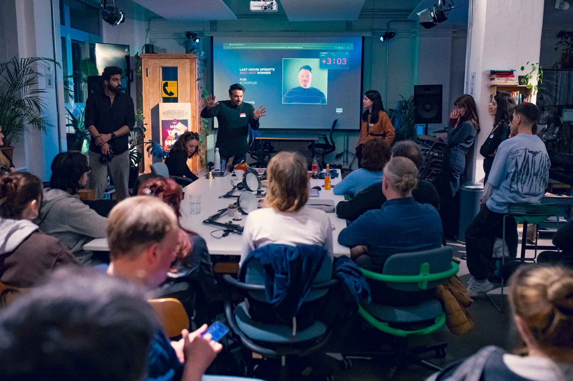 A group of people attending a presentation or workshop in a conference room. A presenter stands at the front, with a large screen behind him showing a photo of a man and some text. Audience members are seated around tables, some taking notes or looking at their devices.