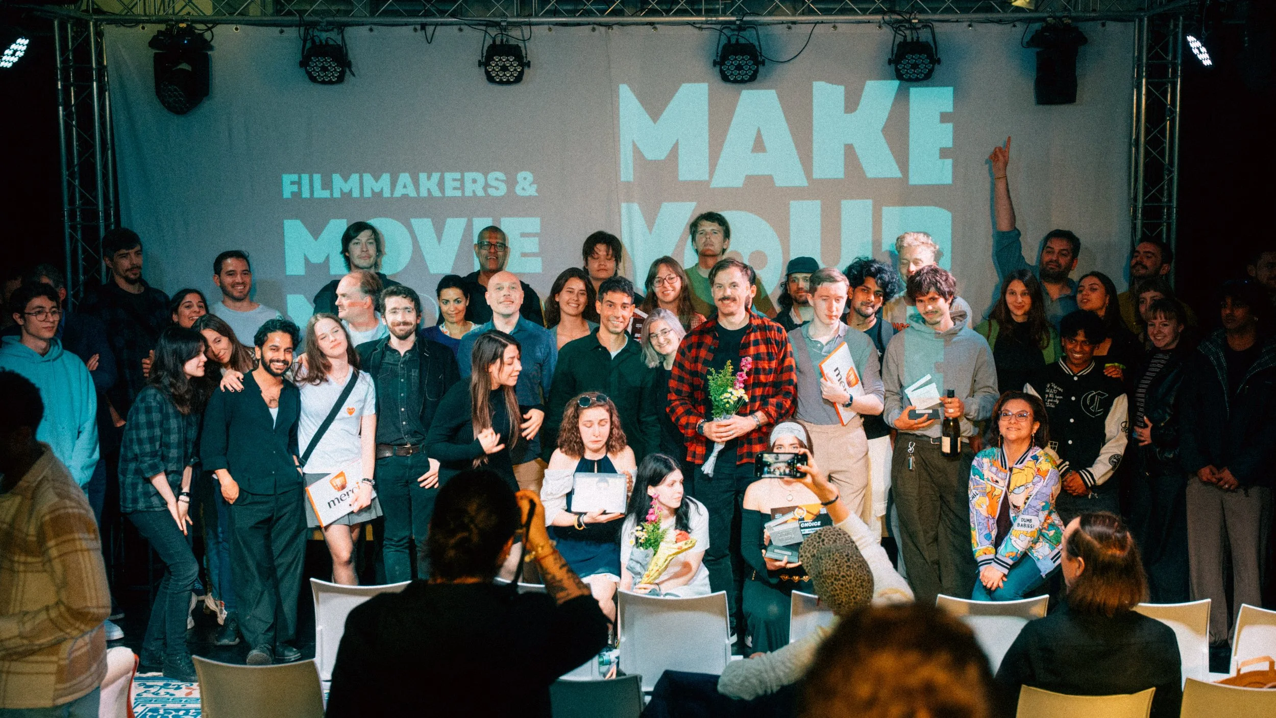 A large group of diverse people on stage at a film event, with a backdrop that reads 'Make Move' and 'Filmmakers & Move' in bold blue and white text. One person in the front holds a bouquet of flowers, others hold certificates or bags, and many are smiling or looking towards the audience.