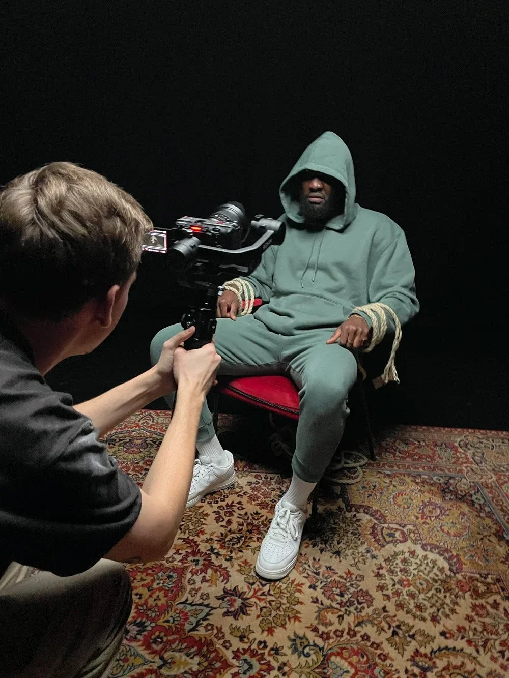 A man in a green hoodie with the hood up sits on a chair on a patterned rug, while another person records him with a camera.
