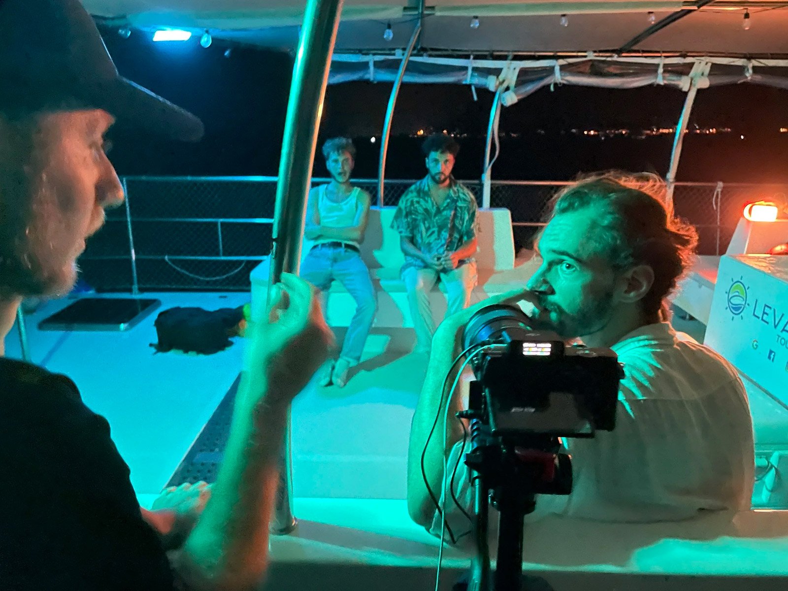 Group of people on a boat at night, illuminated by colored lights, with some sitting on a bench and others engaging in conversation or taking photos.
