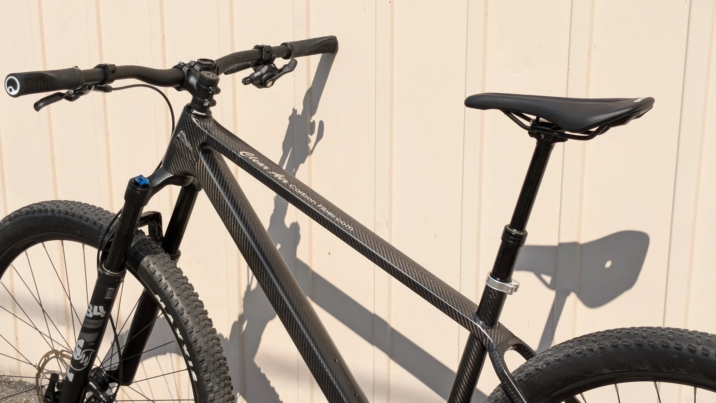 Close-up of a black carbon fiber mountain bike with thick tires and a suspension fork, leaning against a white wooden wall.