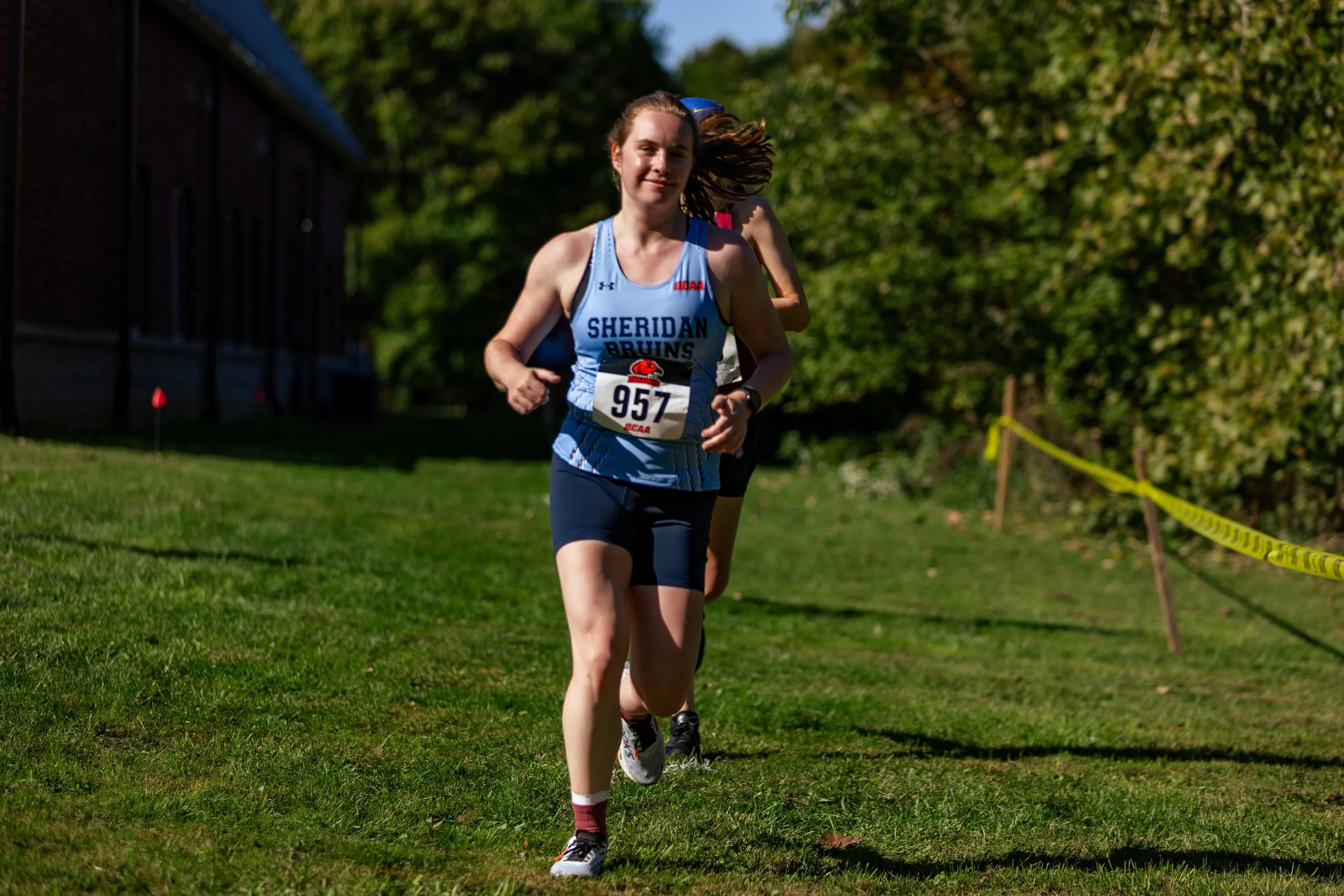 A young woman running in a park during a cross-country race, wearing a blue and white Sheridan Bruins jersey with the number 957, on a grassy course with trees and a yellow rope marking the path.