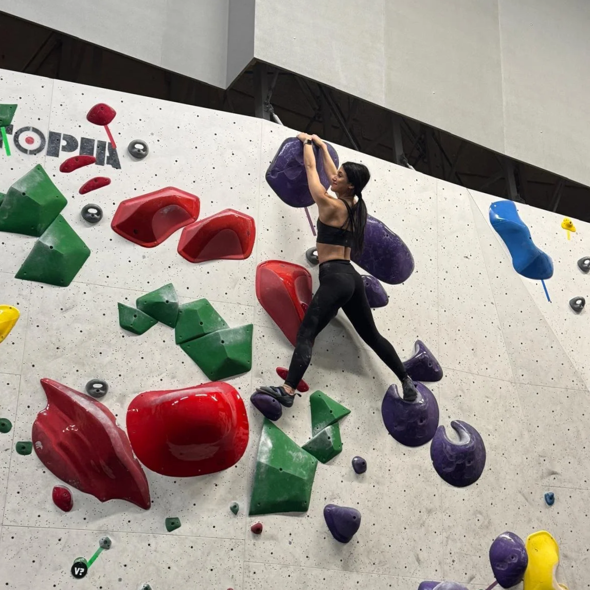 A woman climbing an indoor bouldering wall with multicolored holds, wearing a black sports bra and leggings.