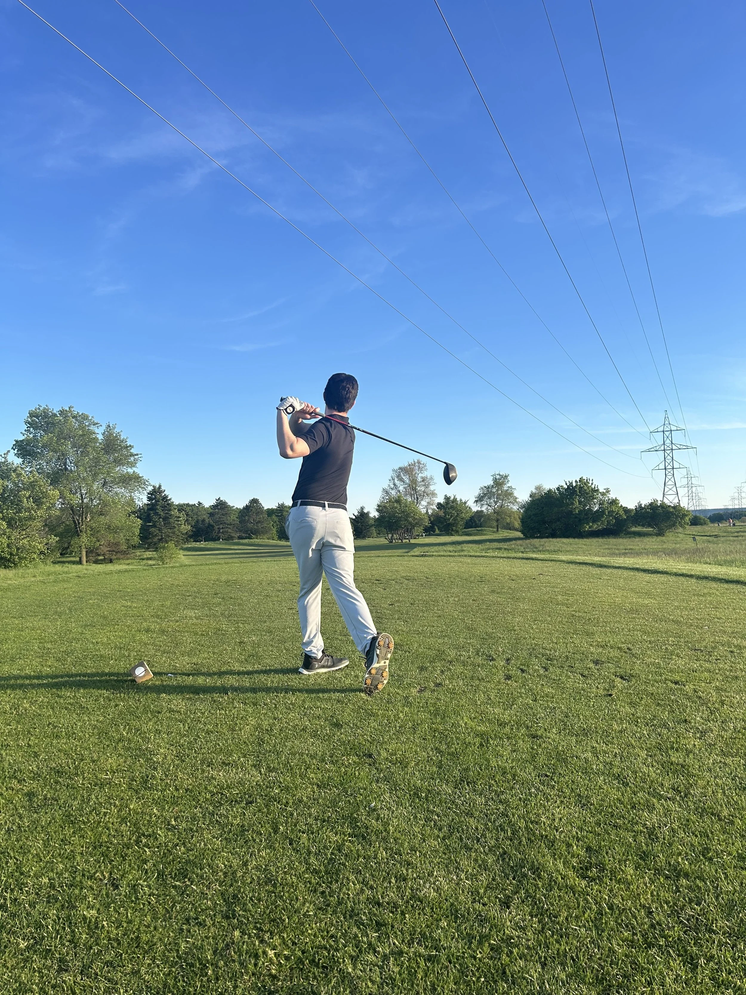 A man playing golf on a green golf course with power lines and trees in the background under a clear blue sky.