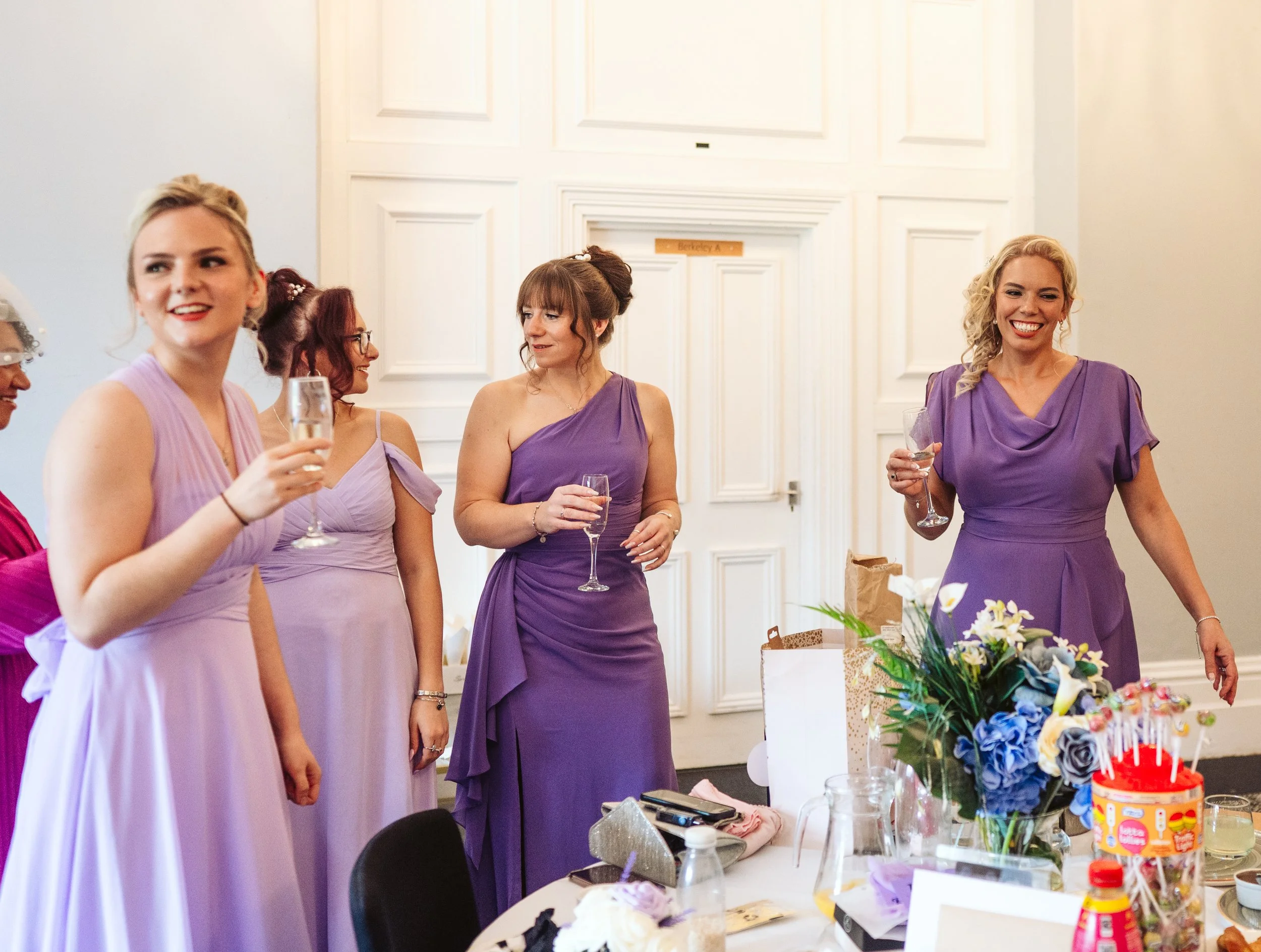 Women in purple dresses holding glasses at a celebration or wedding reception in a decorated room.
