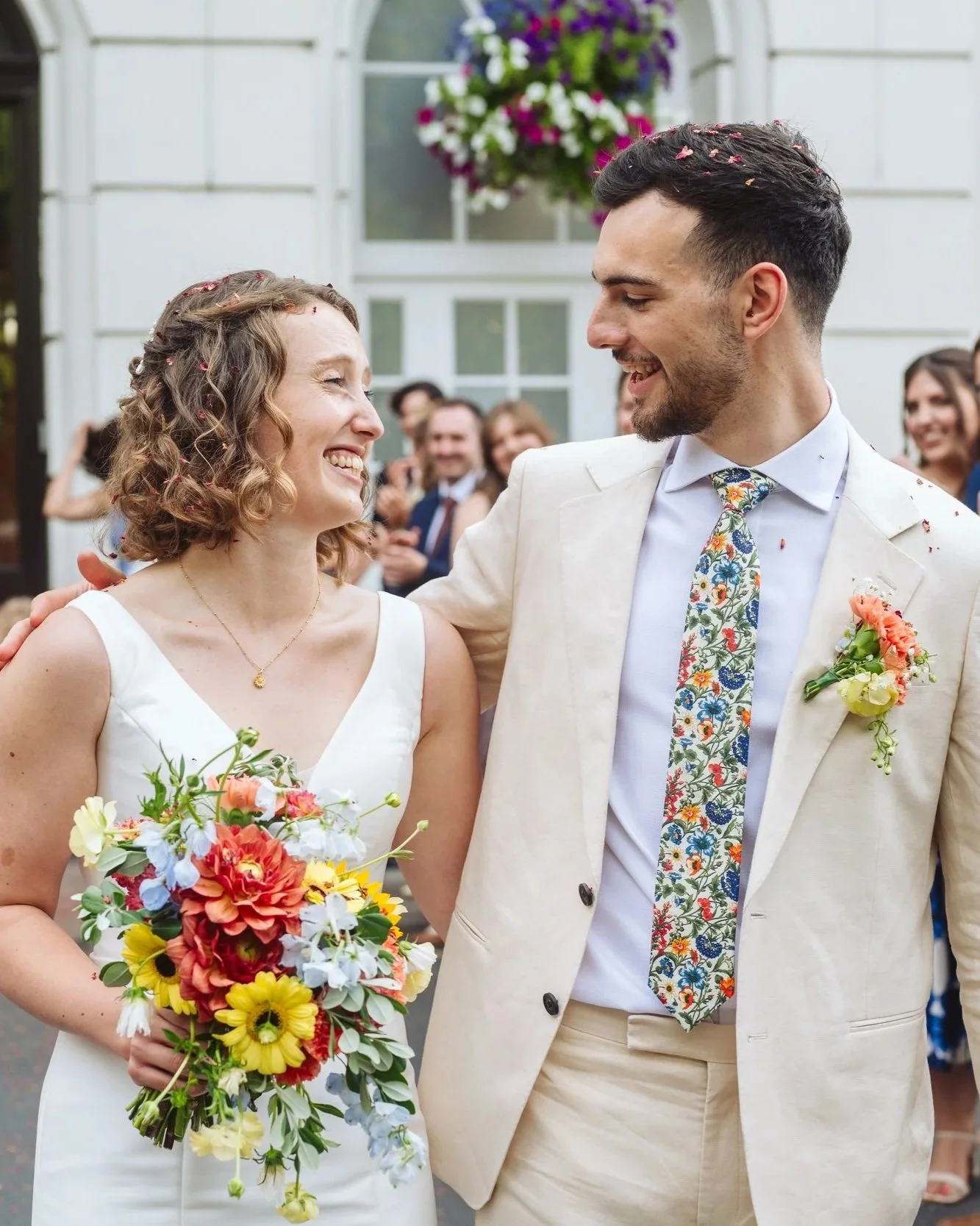 A bride and groom smiling at each other during their wedding, with the bride holding a colorful bouquet of flowers, and wedding guests in the background.