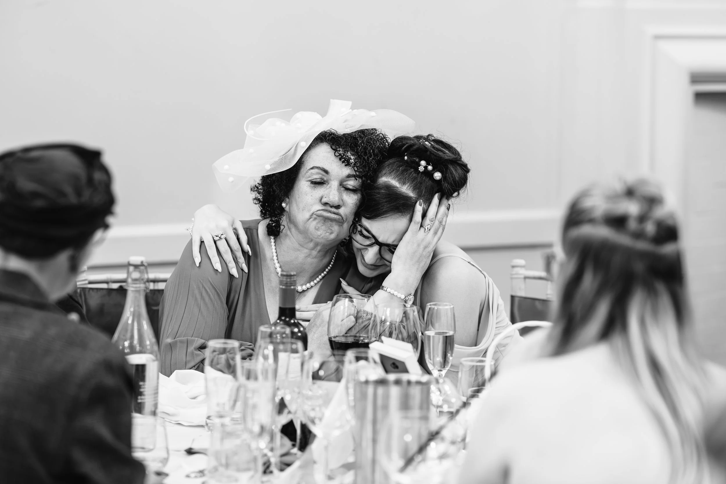 Two women at a wedding reception hugging and comforting each other. One is wearing a hat with a veil and the other has glasses and hair accessories.