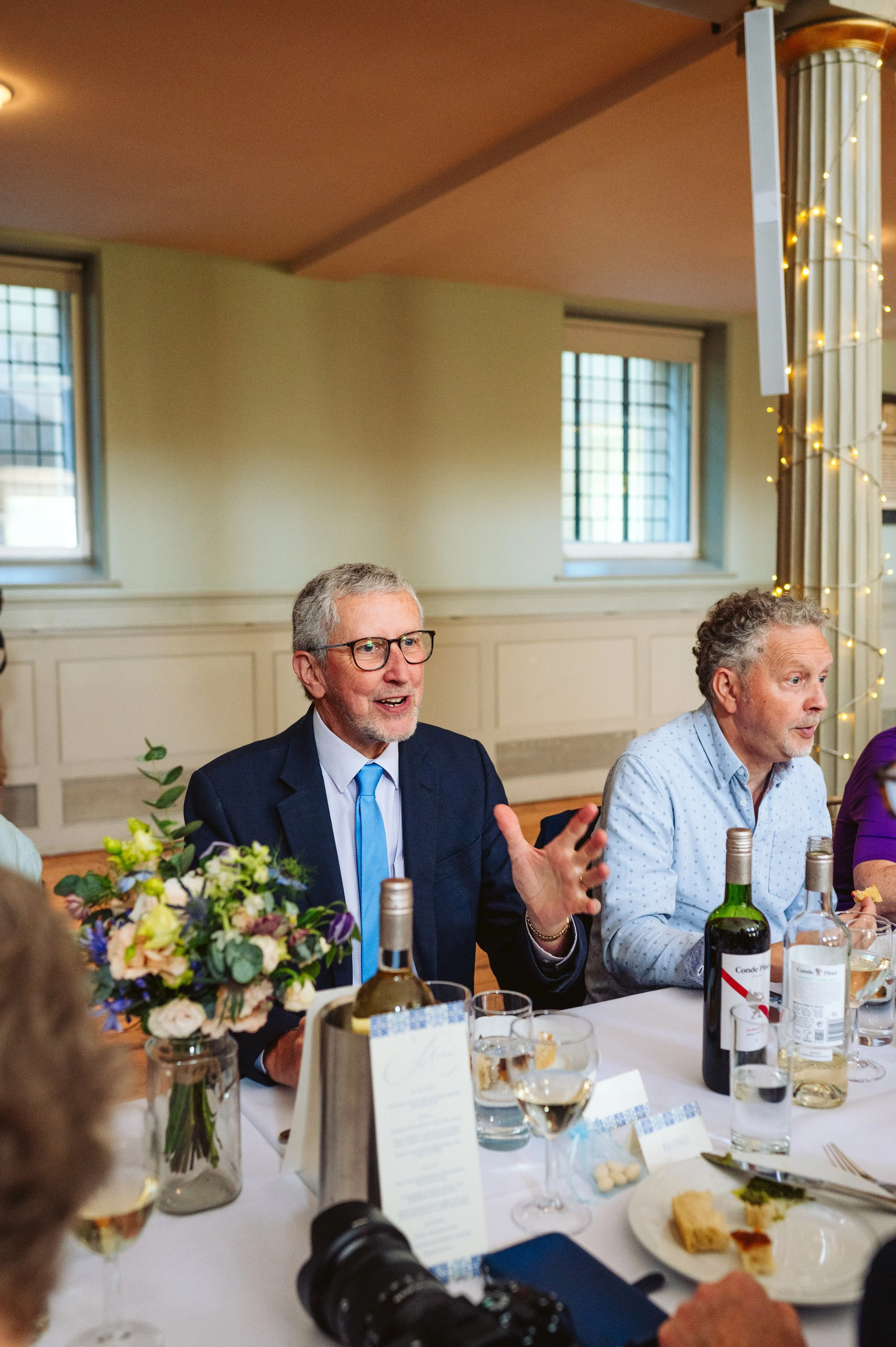 Man in a dark suit and glasses speaking at a formal gathering, sitting at a table with flowers, wine, and food, with another man beside him.