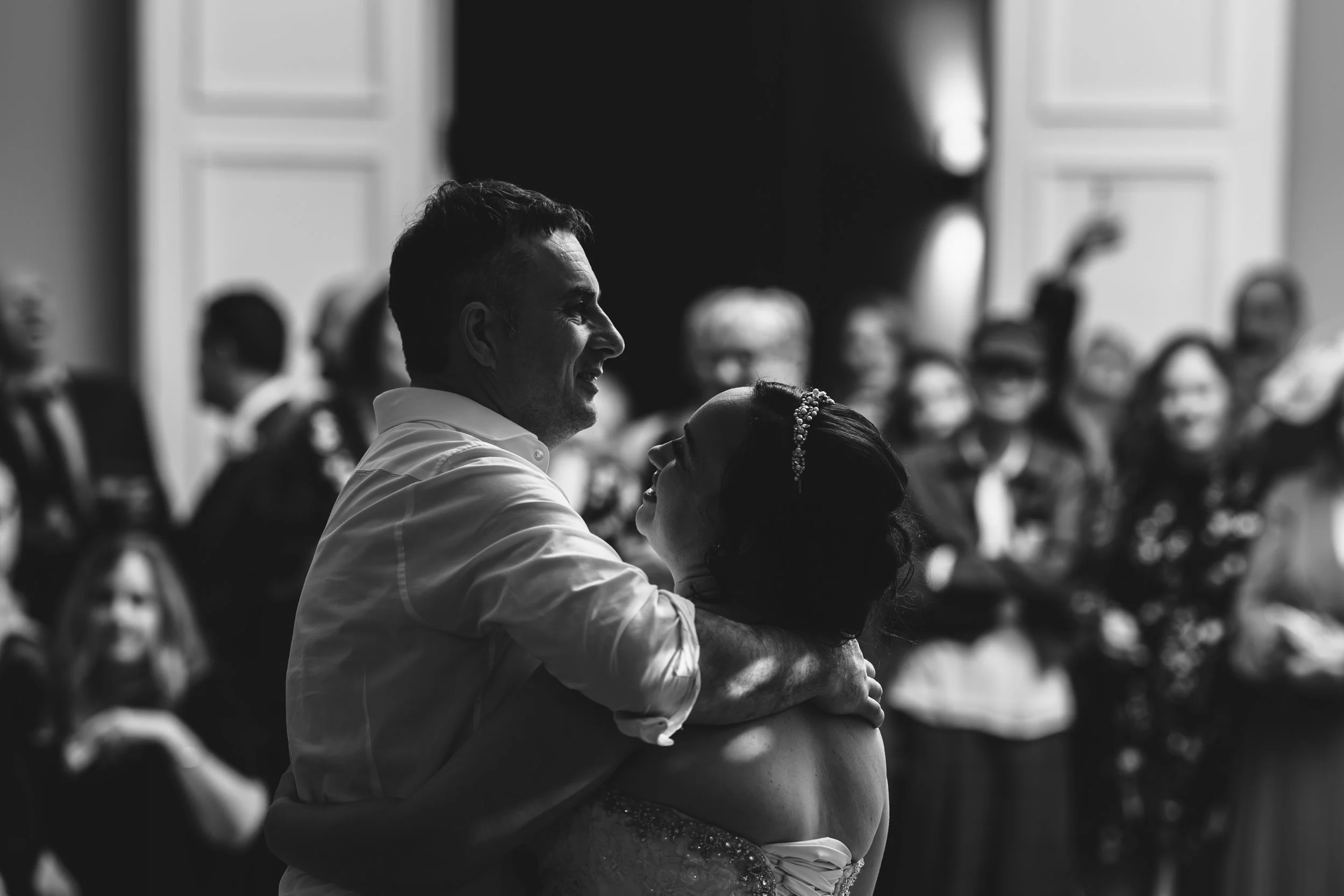 Black and white photo of a couple dancing closely, surrounded by guests at a wedding reception.