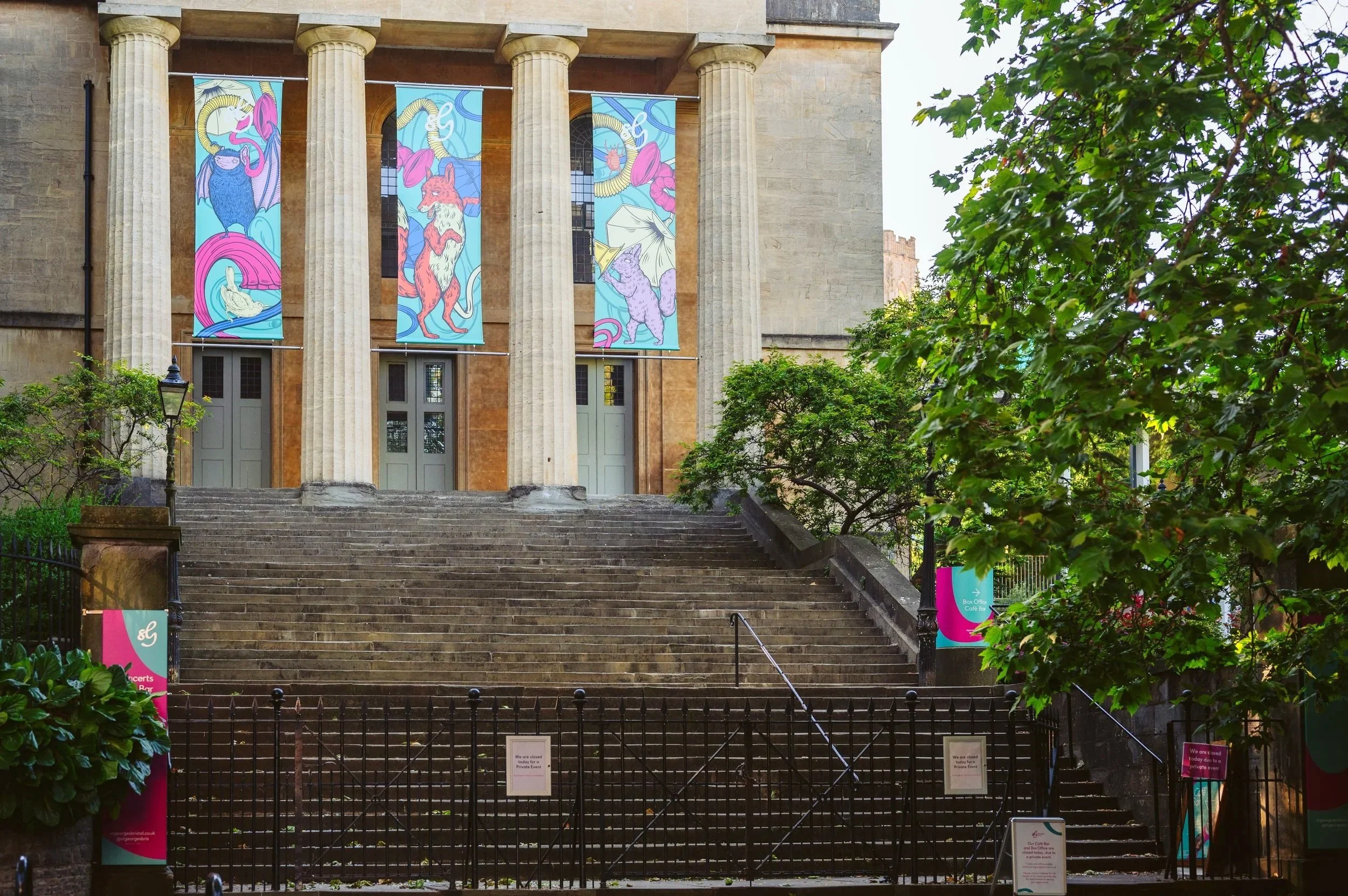 The front of a historic building with four large, fluted columns and stairs leading up to the entrance. The building has three large, arched windows and colorful banners with artwork of mythical creatures hanging between the columns.