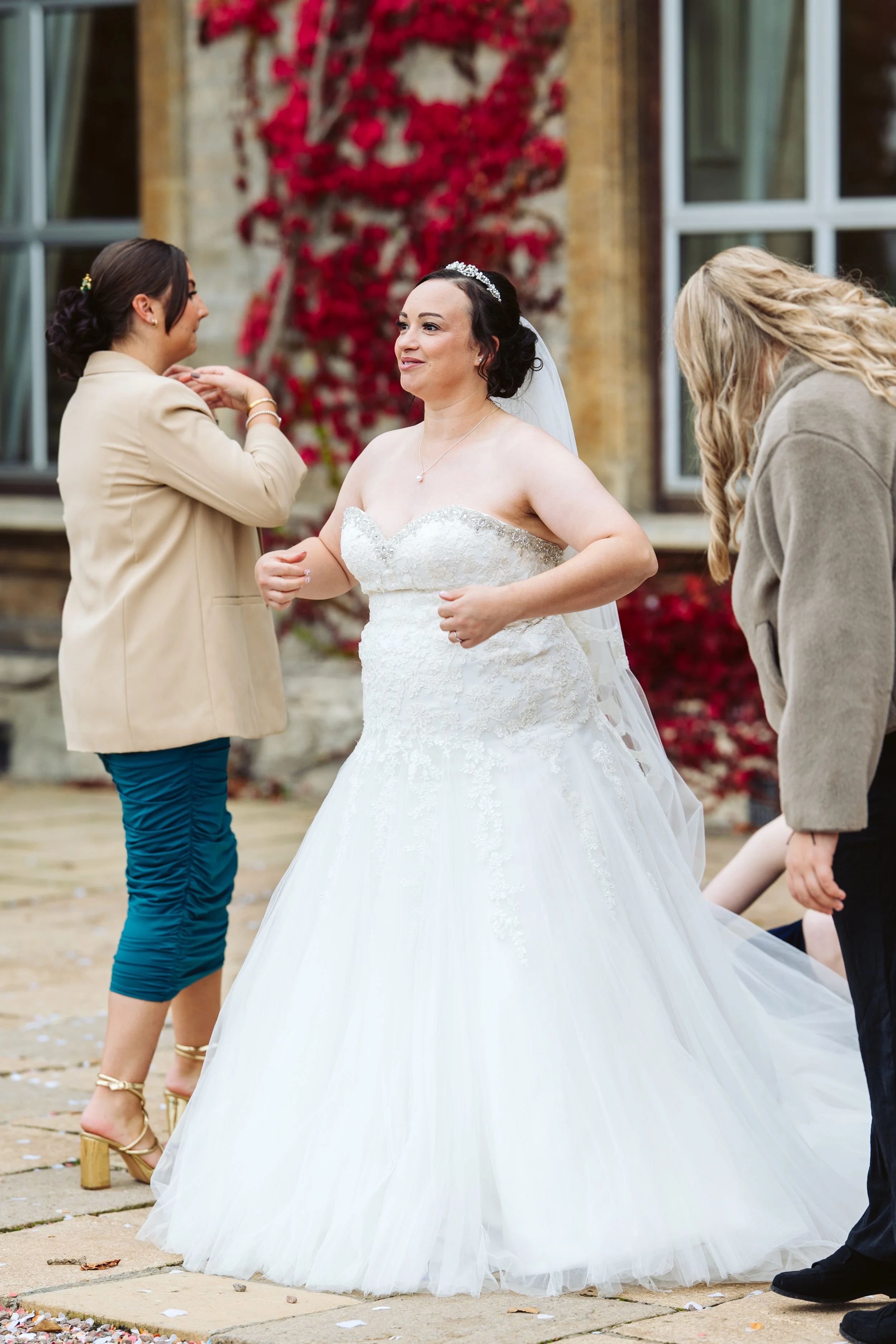 A bride in a white wedding dress with a tiara and veil standing outdoors, with two women preparing her for the wedding.