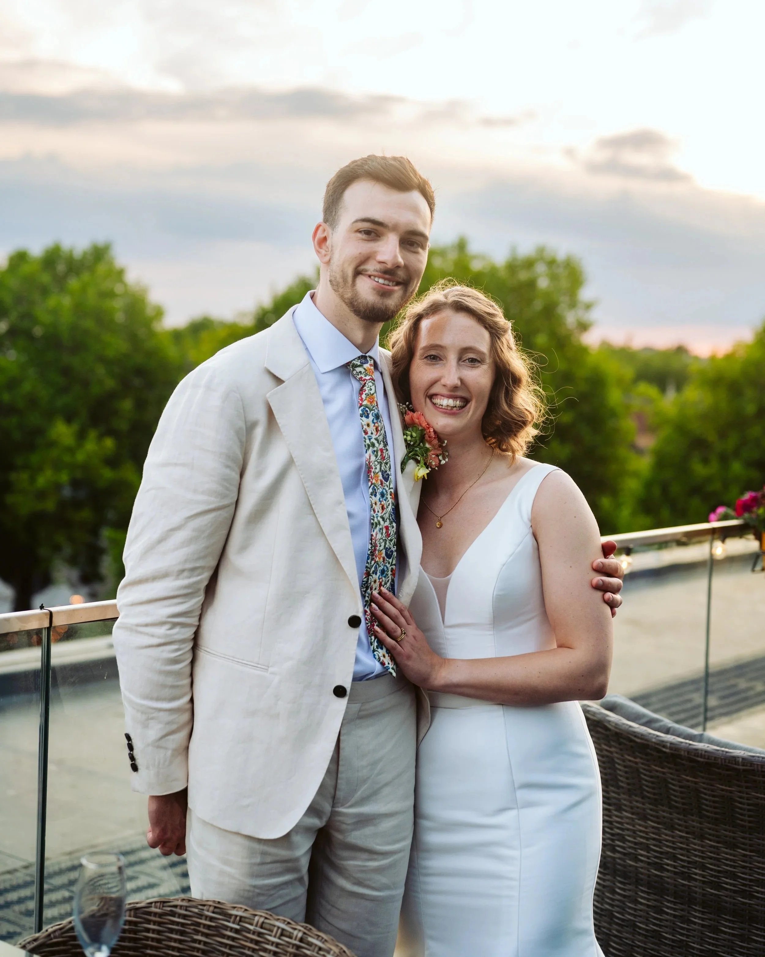 A recently married couple smiling and posing outdoors on a balcony during sunset. The man wears a cream suit with a floral tie, and the woman wears a white wedding dress with a floral boutonniere.