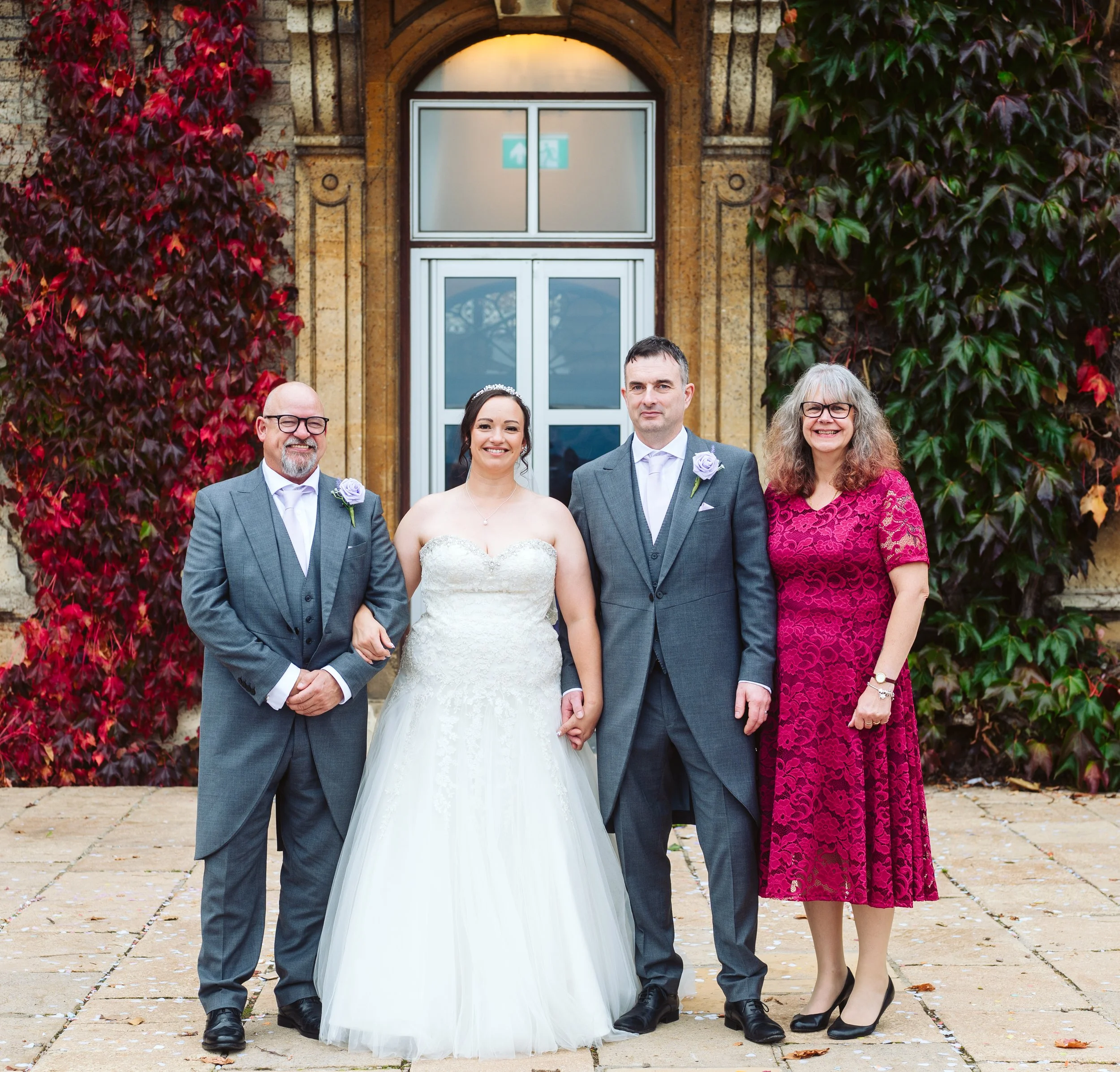 A wedding photograph of a couple with two older adults standing outdoors in front of a stone building with an arched window and red and green ivy. The bride wears a white wedding gown, and the groom and the man to his left wear gray suits with lavender boutonnières. The woman on the far right wears a pink lace dress.