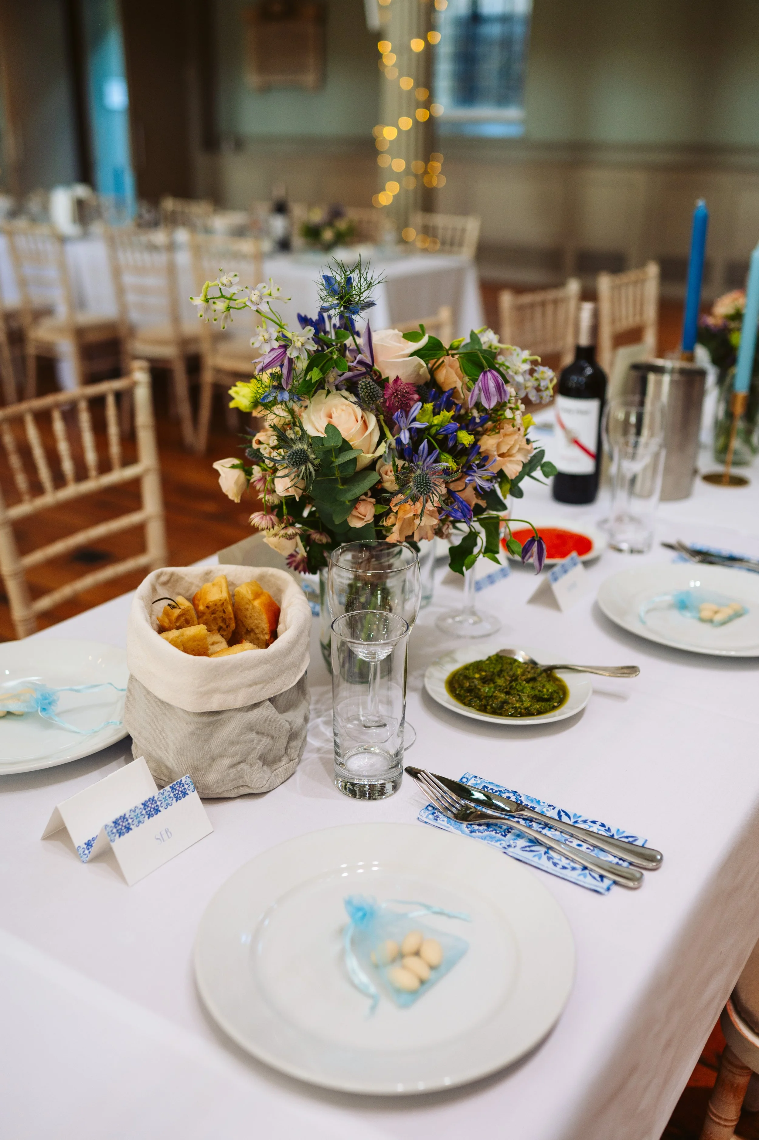 A decorated table at a formal event with a floral centerpiece, bread in a cloth bag, various plates, glasses, cutlery, and a bottle of wine.