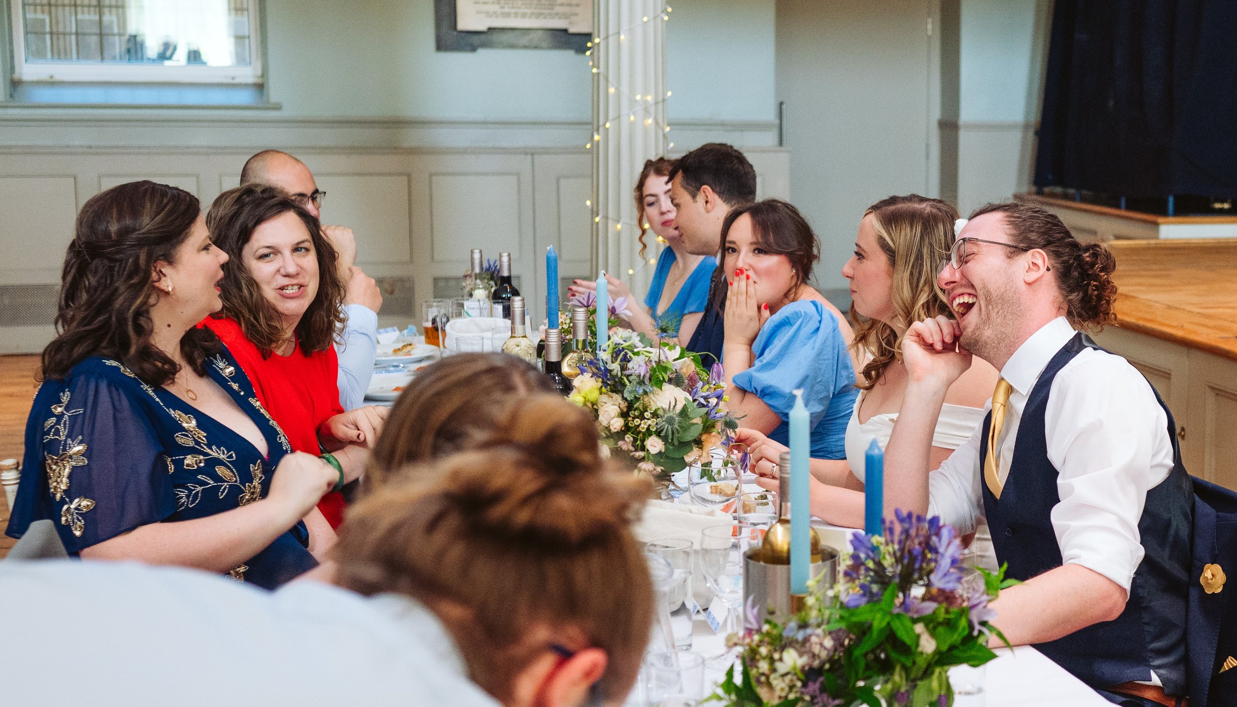 People sitting at a long dining table, laughing and talking during a celebration event, with floral centerpieces and blue candles.