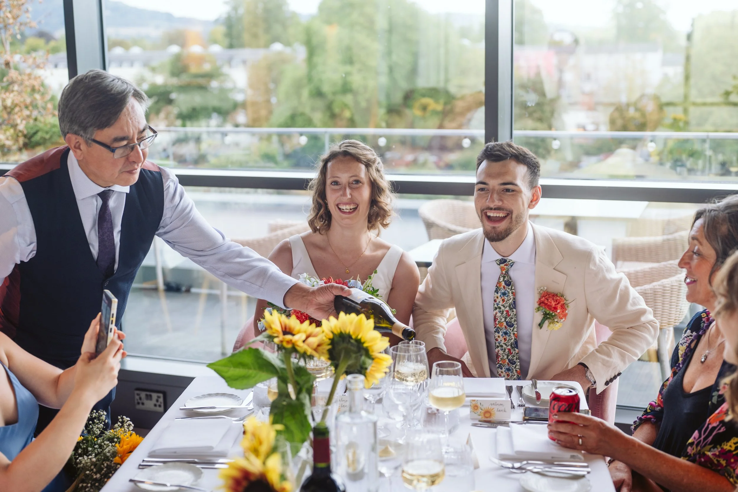 People celebrating at a wedding reception, with a waiter pouring wine. The group is sitting at a table decorated with sunflowers, and they are smiling, enjoying the moment.