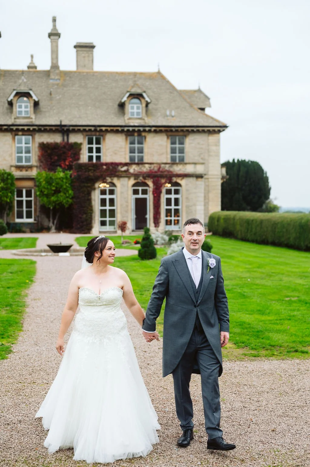 Bride in a white wedding dress and groom in a gray suit walking hand in hand outside a large historic house with green lawn and garden.