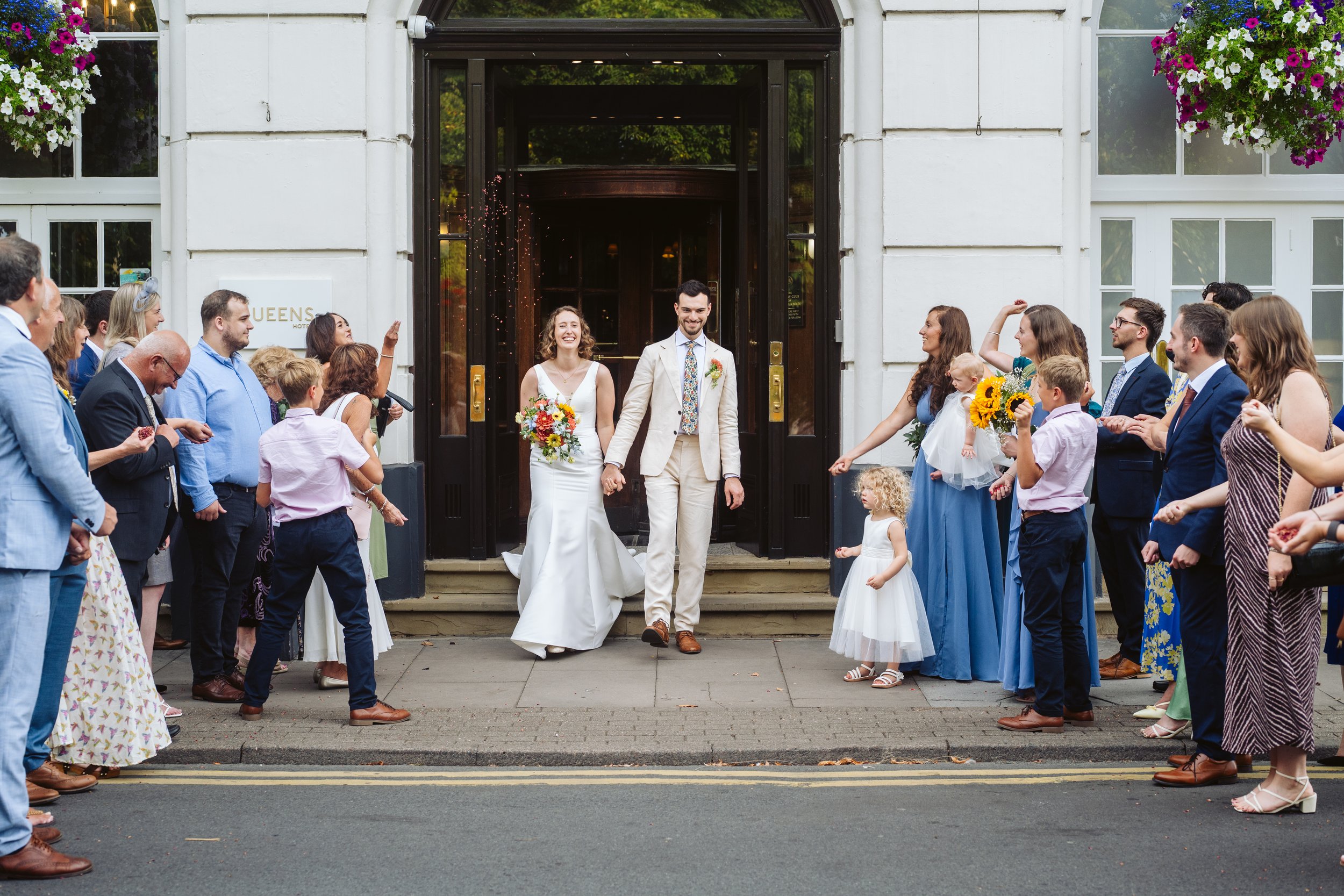 A bride and groom holding hands and smiling as they walk out of a building, surrounded by friends and family celebrating their wedding with cheering and flowers outside a hotel entrance.