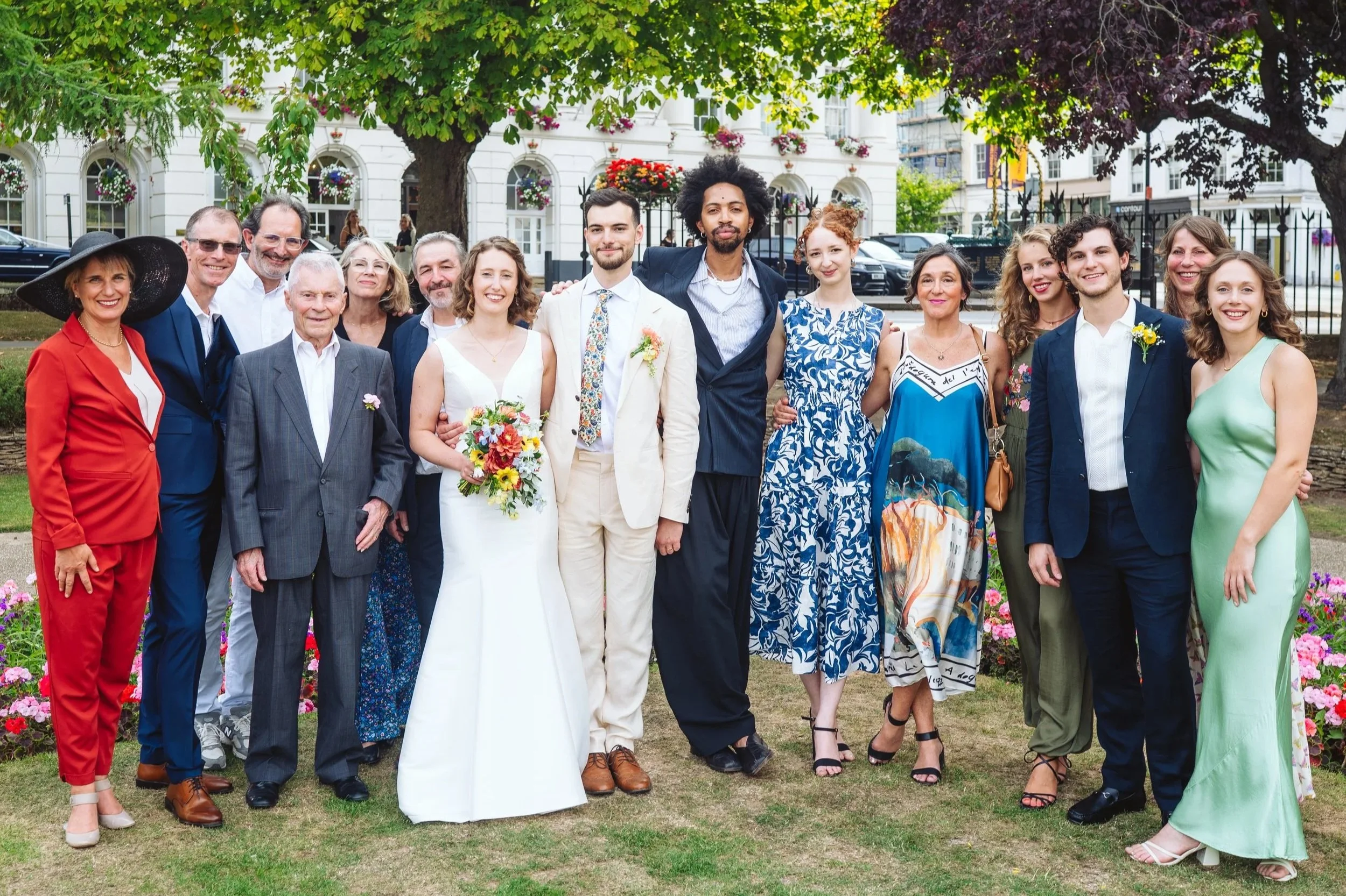 A group of people standing outdoors in a garden during a celebration, with trees, flowers, and a white building in the background. They are dressed in formal attire, including a bride in a white wedding gown holding a bouquet, and others in colorful dresses and suits.