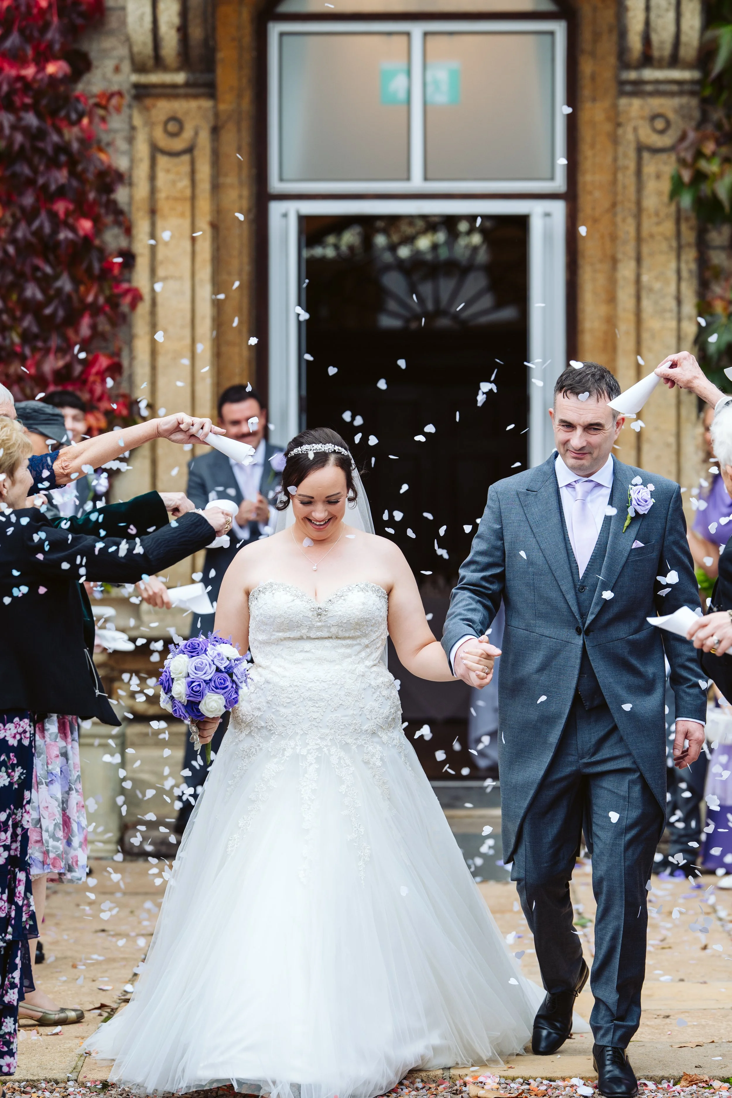 Bride and groom holding hands and walking out of a church, surrounded by guests throwing confetti, with the bride holding a bouquet of purple and white roses, on a fall day.
