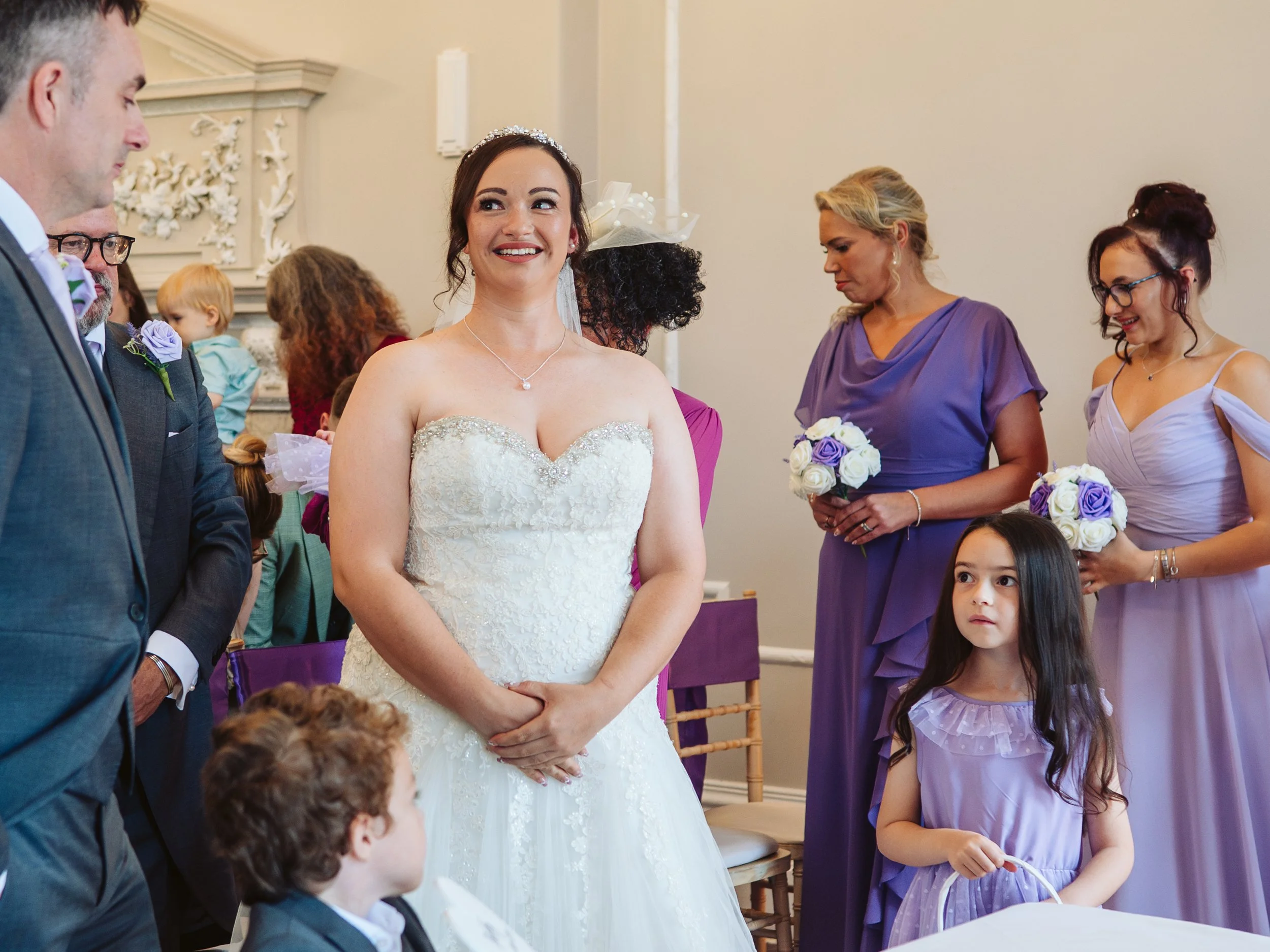 A bride in a white wedding gown smiling during a wedding ceremony, surrounded by bridesmaids in purple dresses holding bouquets, children in the foreground, and guests in the background.