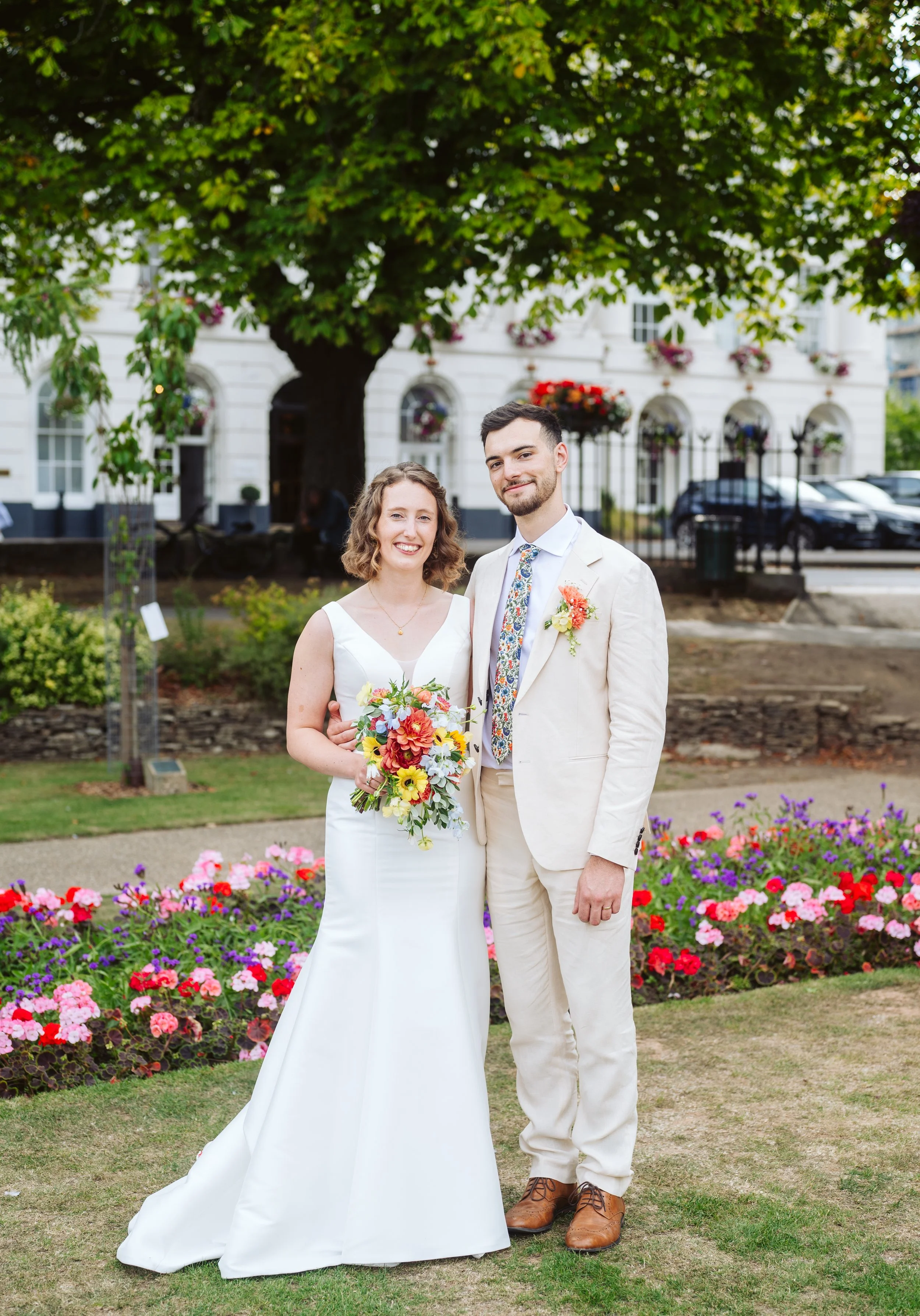 A newlywed couple standing outdoors in a garden with colorful flowers, smiling, with a large tree and white building in the background.