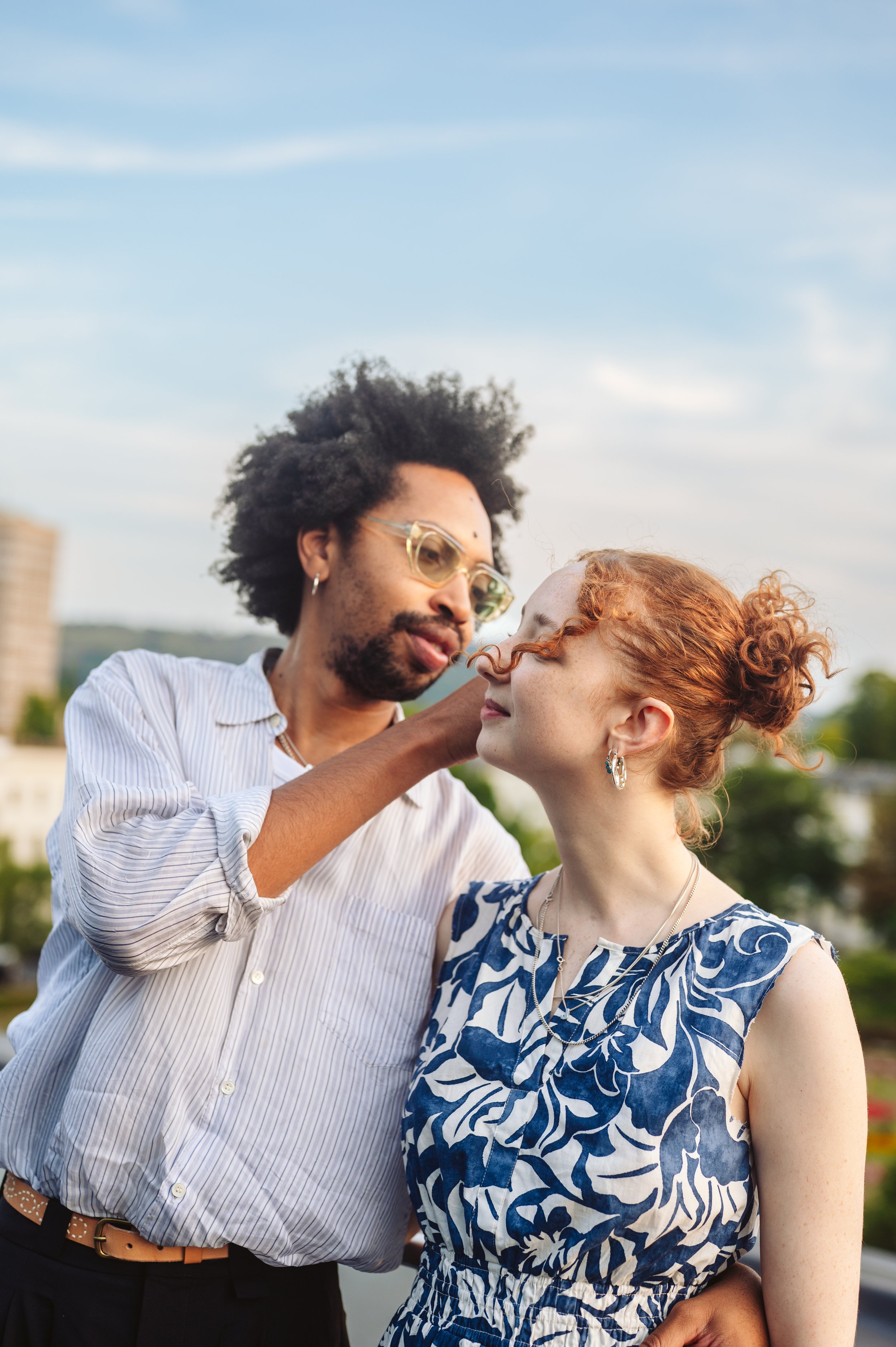 A man with glasses and a white striped shirt adjusts the face of a red-haired woman with earrings and a blue patterned dress, outdoors under a blue sky.