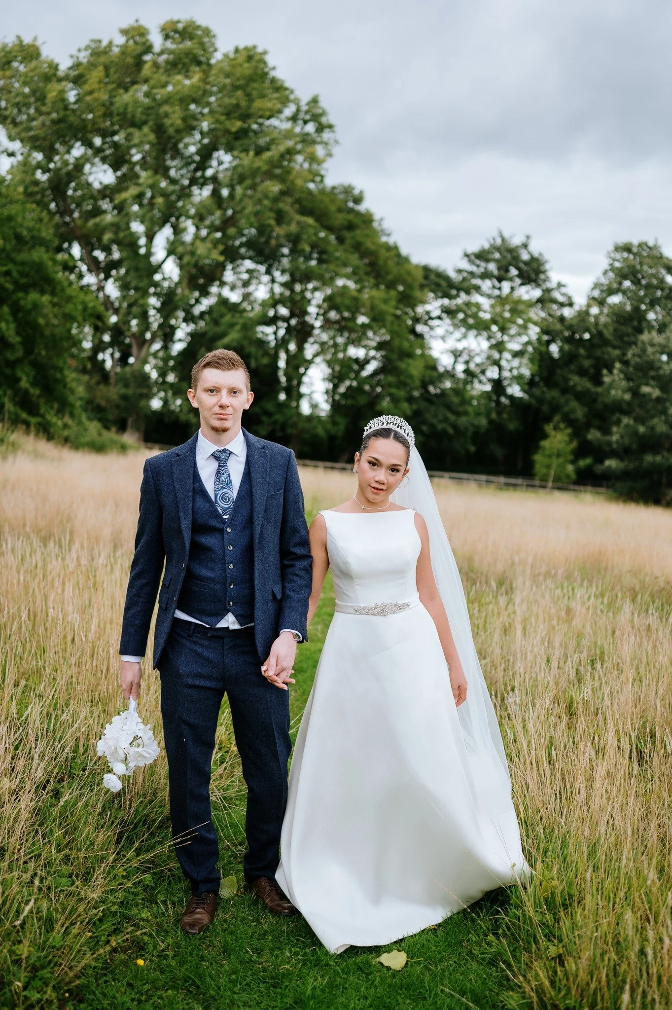 Bride and groom standing hand in hand in a grassy field, with trees and cloudy sky in the background. The bride wears a white wedding gown with a veil and tiara, and the groom wears a dark blue suit and tie, holding a bouquet of white flowers.