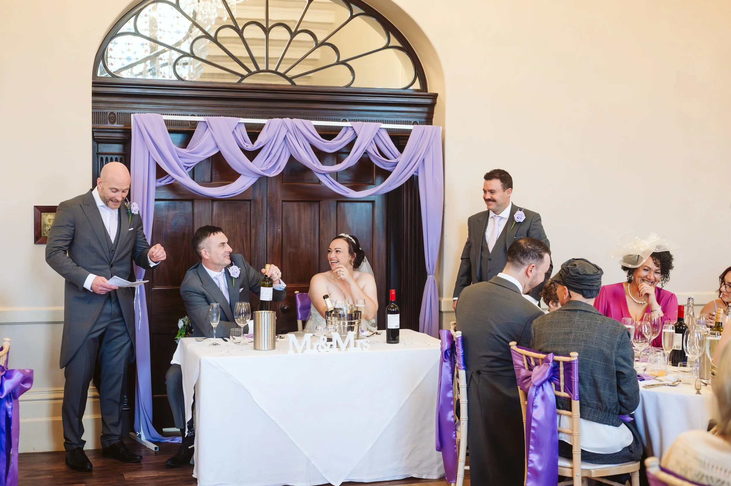 Wedding reception scene with the bride and groom sitting at a table, surrounded by friends and family. The bride is smiling and wearing a white dress, while the groom is holding a glass of wine. The table has wine bottles, glasses, and a table sign that reads 'M&M.' Purple decorations and drapes adorn the background, and guests are dressed formally with some women wearing hats.