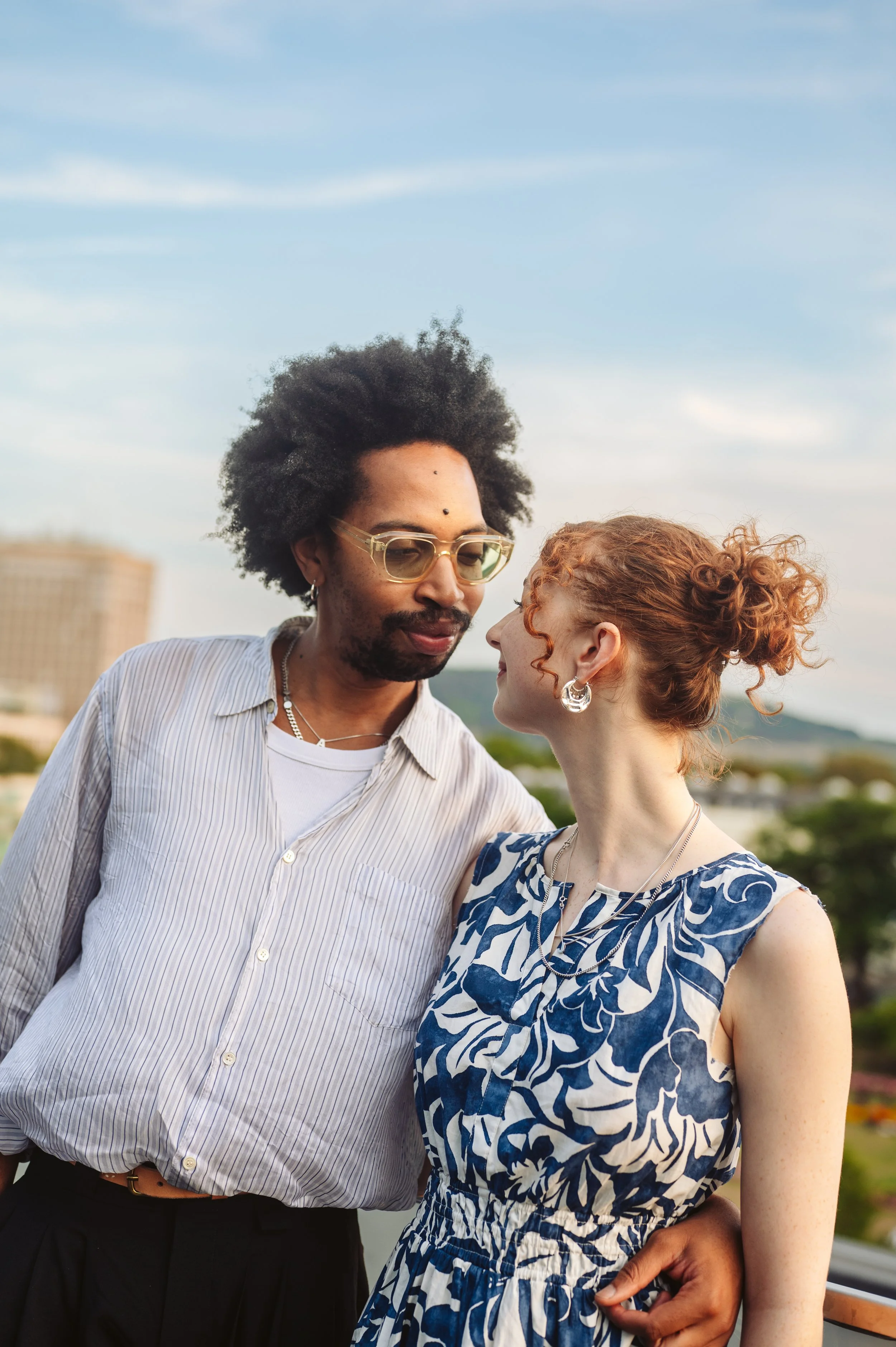 A man and woman stand close together outdoors, looking into each other's eyes, with a cityscape and blue sky in the background.