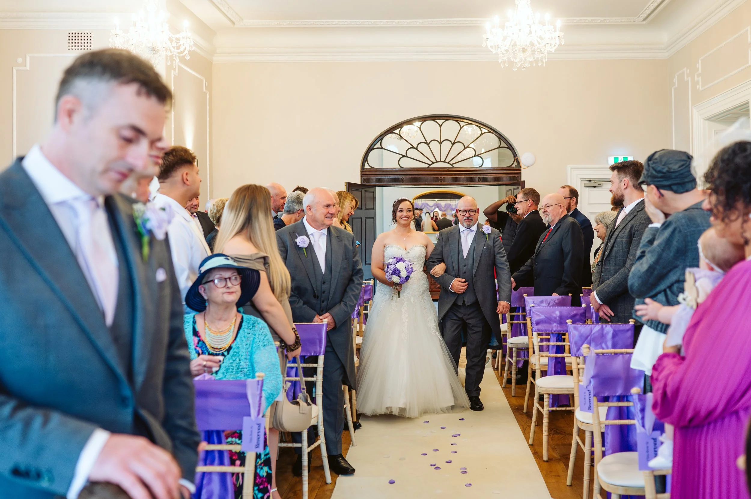 A bride walking down the aisle with her father at a wedding ceremony. Guests are seated on both sides, some standing and watching, with purple decorations and ribbons on chairs.