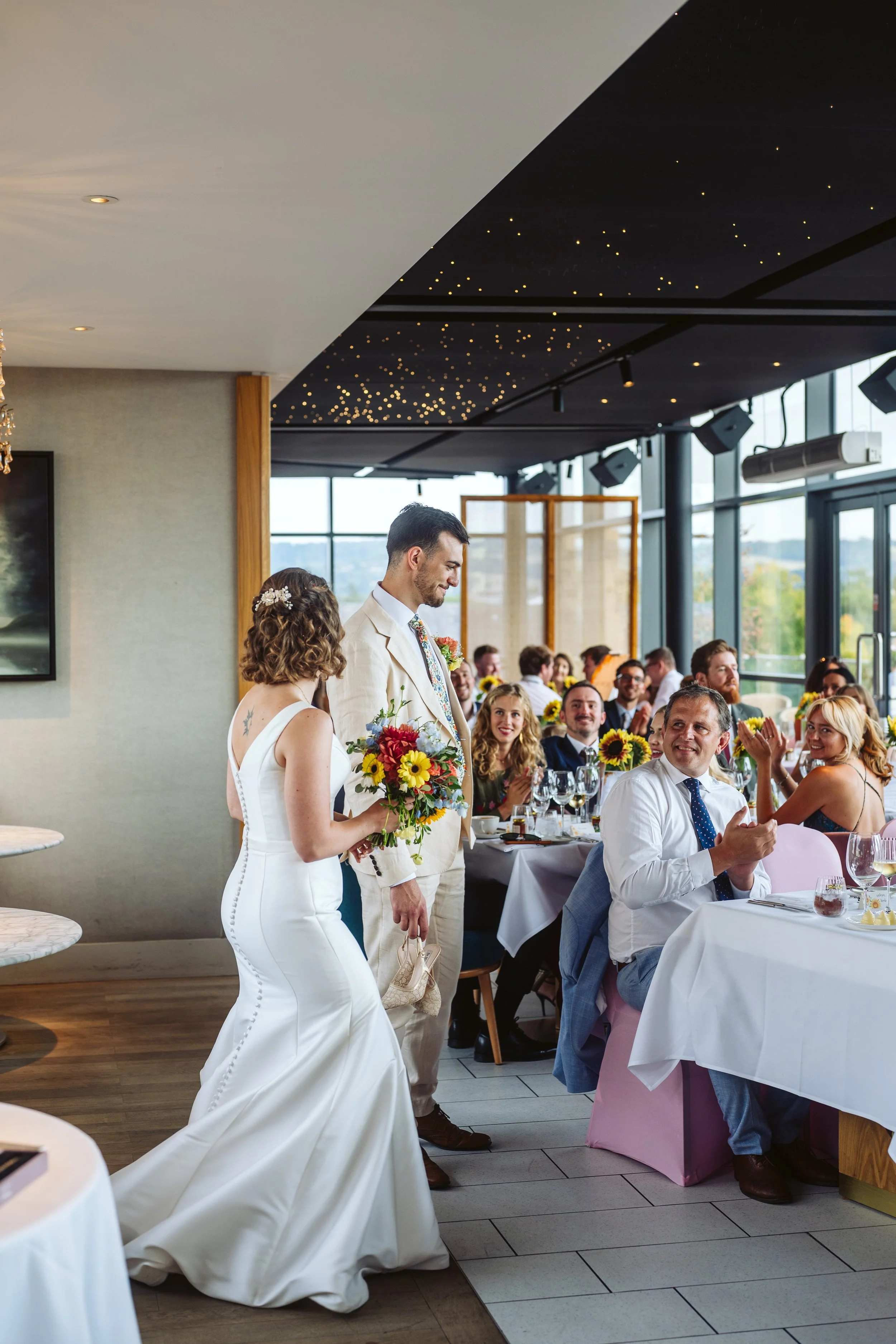 Bride and groom walking into a wedding reception with seated guests clapping and smiling, decorated with sunflowers.