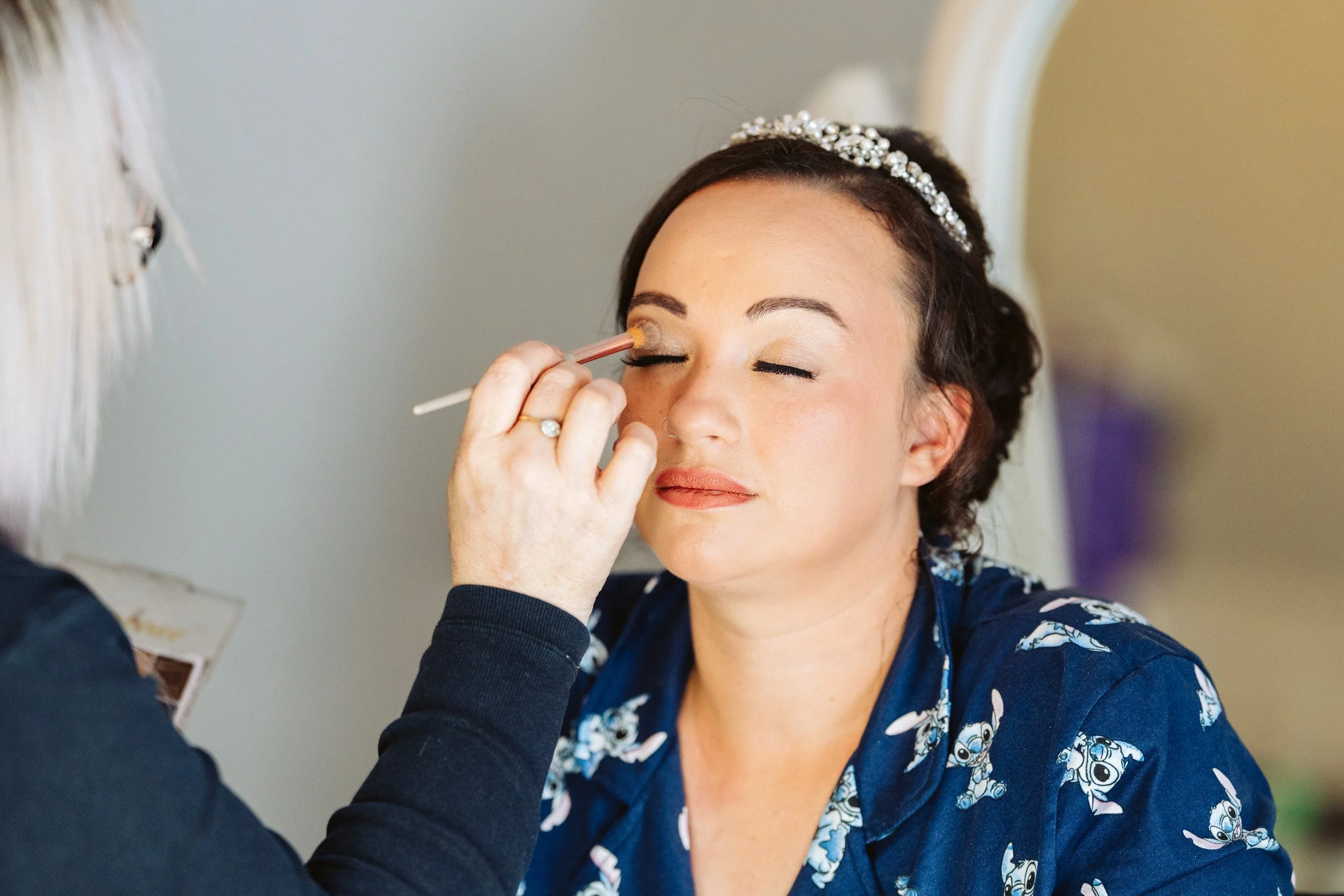 A woman with dark hair and a tiara has her makeup professionally applied, with her eyes closed, while another person applies eyeshadow with a brush.