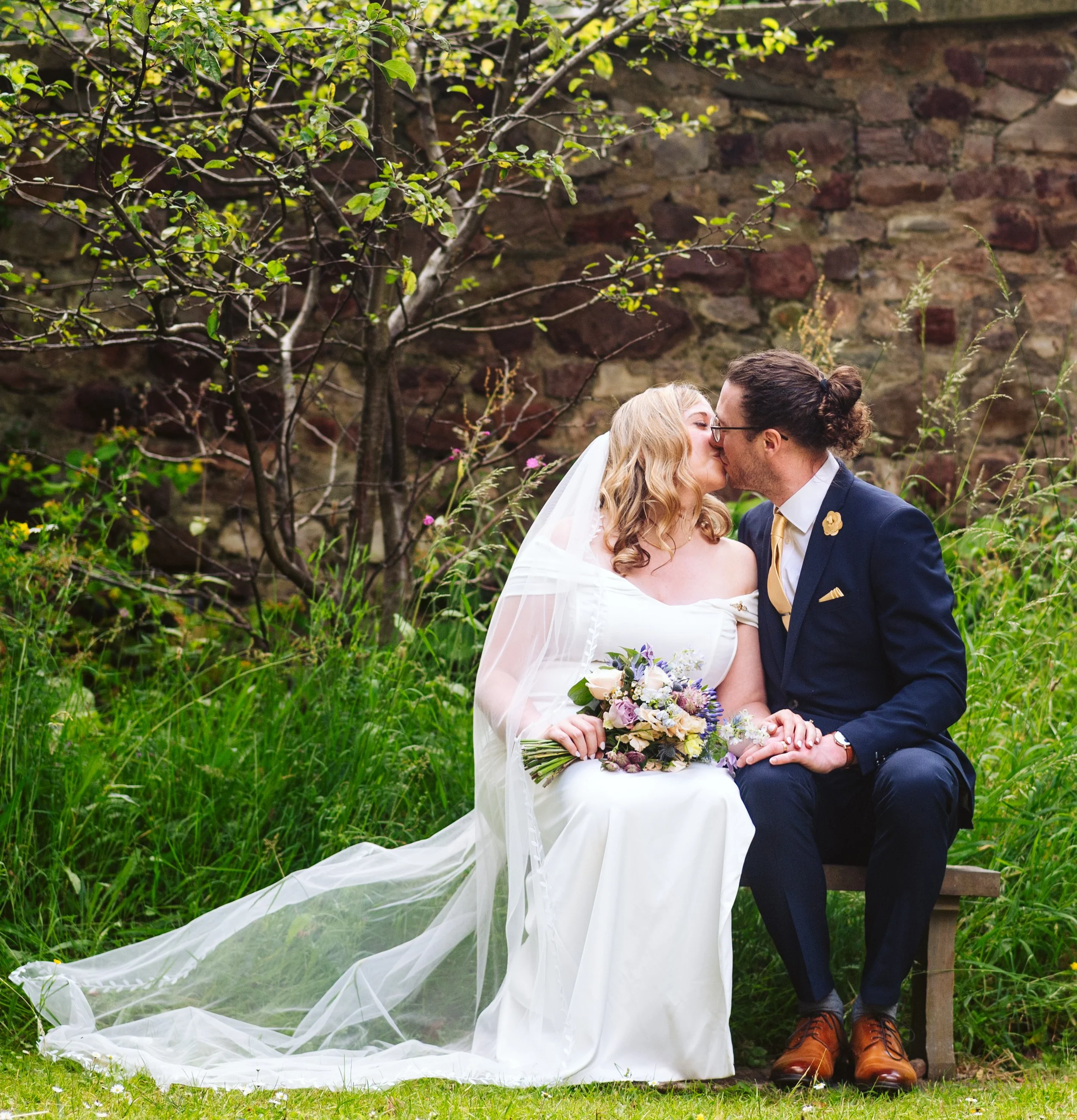 A newlywed couple sharing a kiss on a bench outdoors, surrounded by greenery and a stone wall, with the bride in a white wedding dress and veil holding a bouquet, and the groom in a navy suit with a gold tie.
